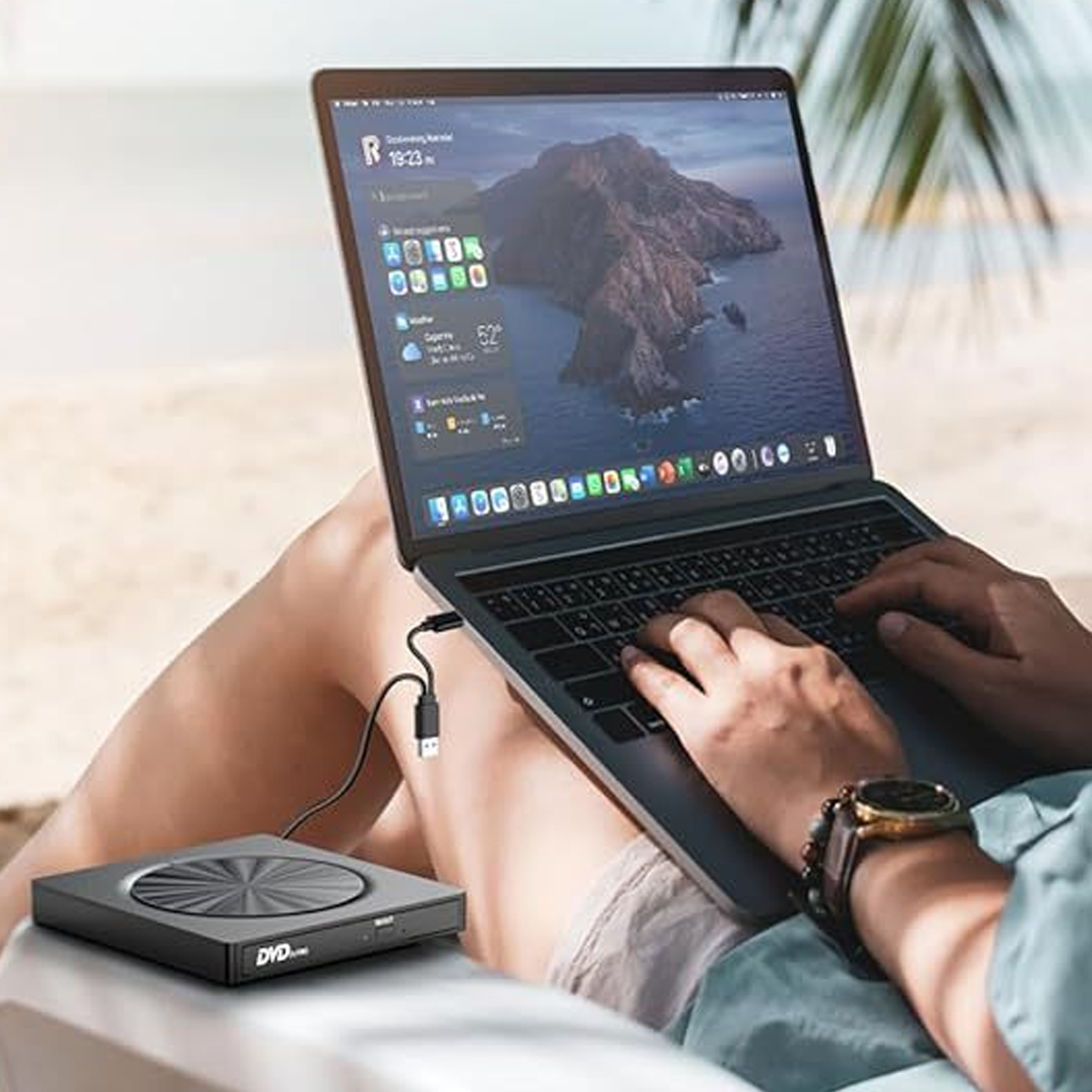 Eine Person nutzt einen Laptop im Freien am Strand, während ein externes DVD-Laufwerk per Kabel verbunden ist.