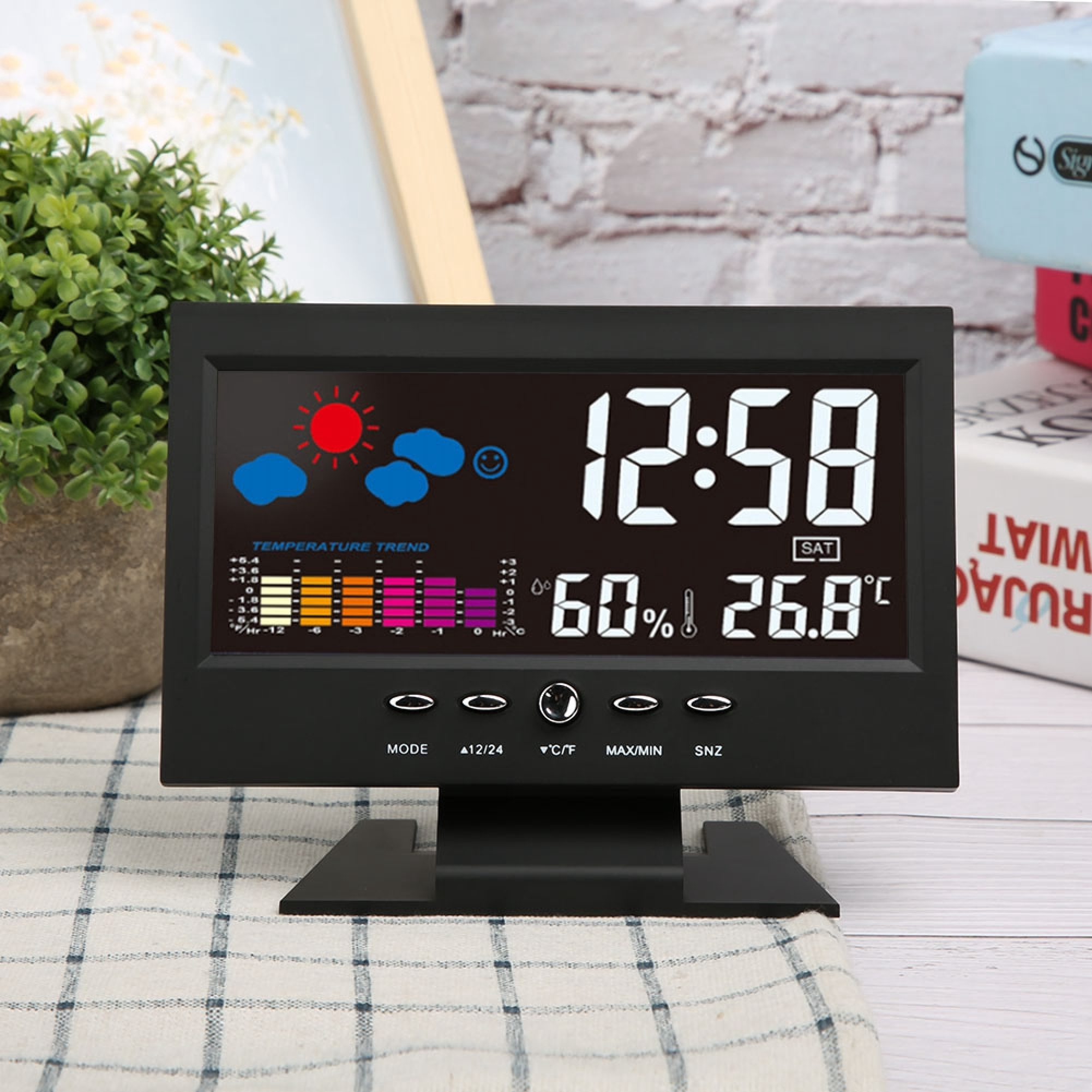 Schwarze digitale Wetterstation mit Uhrzeit, Temperatur, Luftfeuchtigkeit und Wettergrafik auf kariertem Tuch.