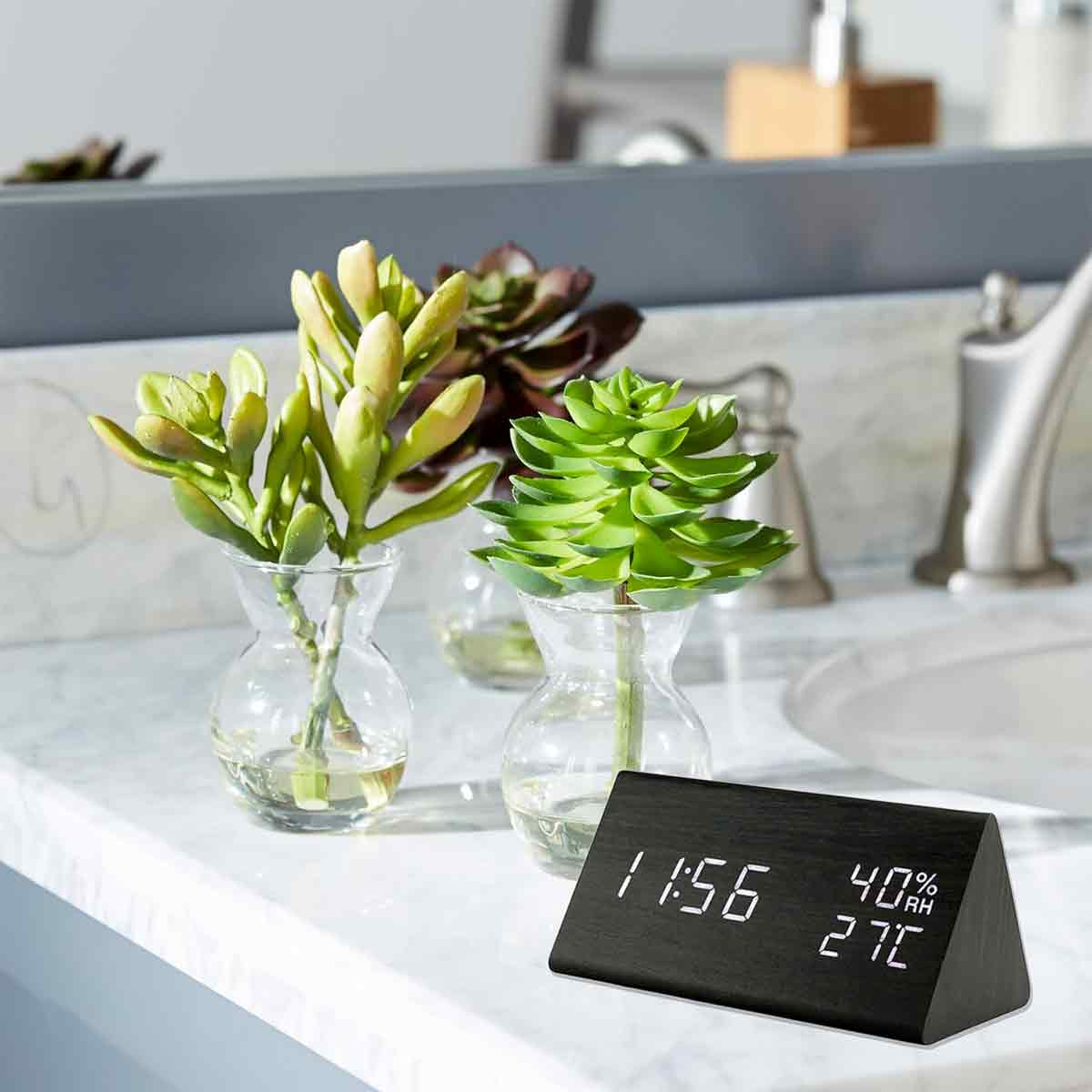 Holzuhr mit digitalem Display, kleinen Sukkulenten in Glasvasen und Waschbecken im Hintergrund.
