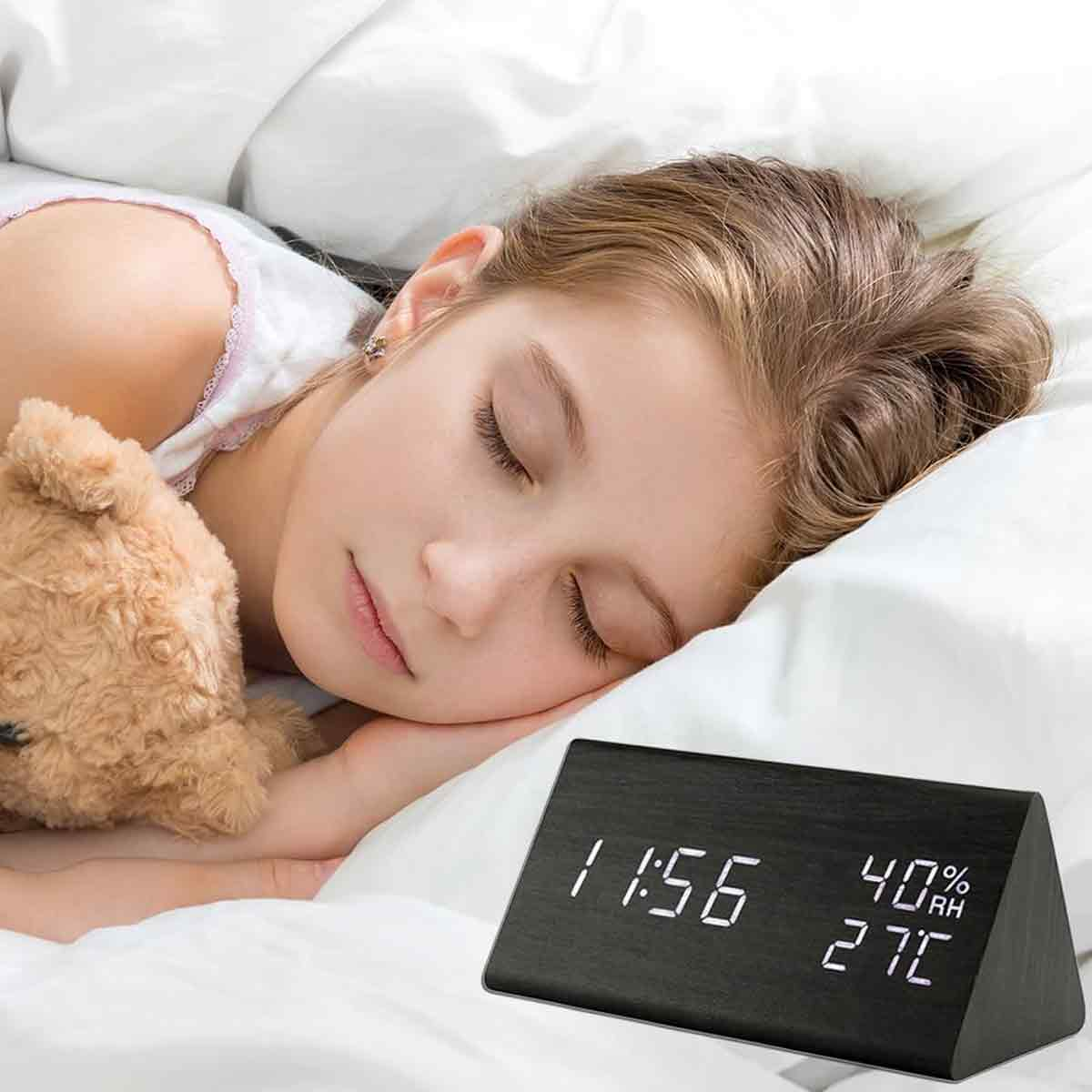Ein junges Mädchen schläft friedlich im Bett neben einem Teddybären, während ein digitaler Wecker 11:56 anzeigt.