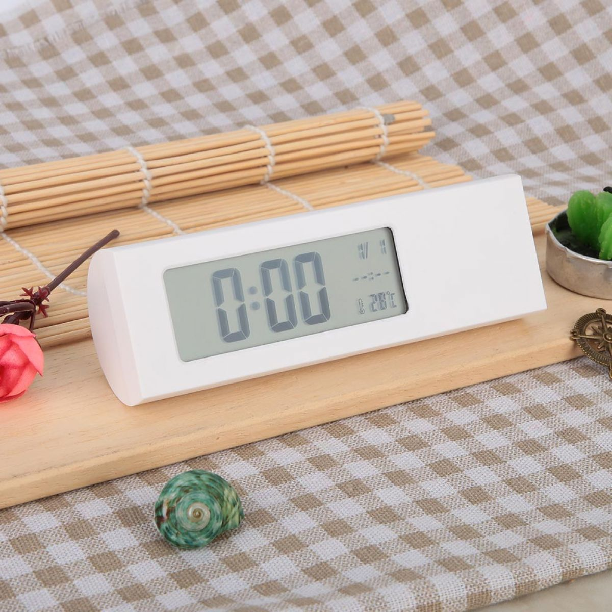Eine weiße Digitaluhr auf einem Holzbrett vor kariertem Hintergrund mit einer Muschel.