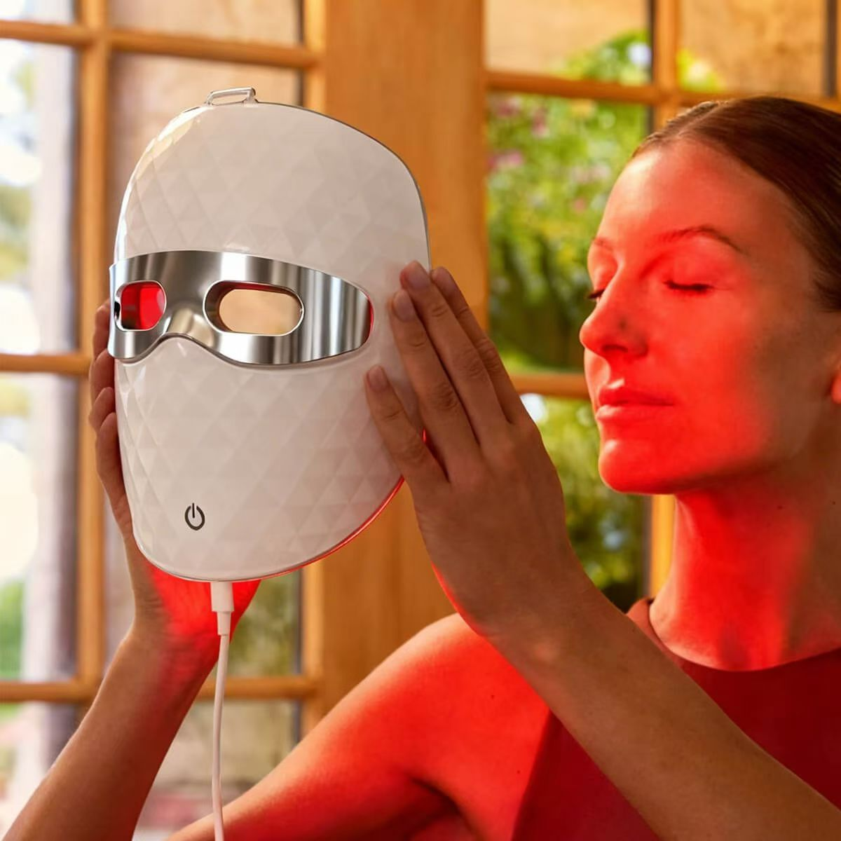 Eine Frau hält eine weiße LED-Lichttherapiemaske mit roten Leuchten vor ihr Gesicht.