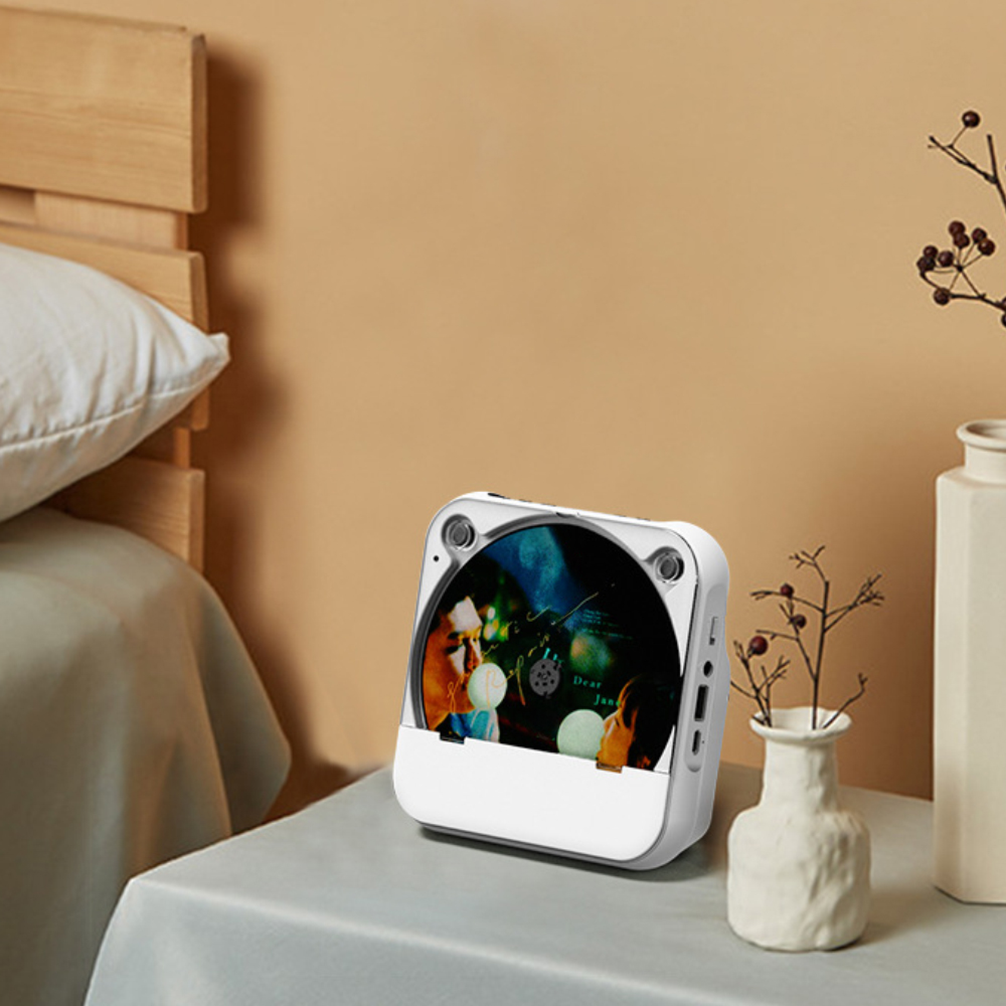 Ein weißer tragbarer CD-Player auf einem Nachttisch mit einer Vase und einem Bett an einer beigen Wand.