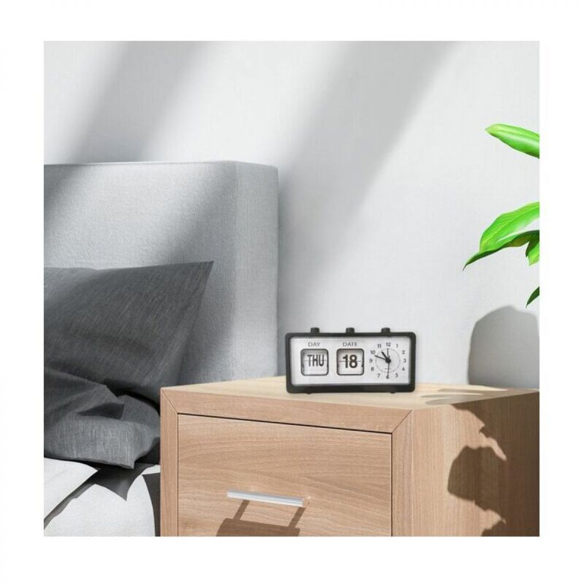 Ein schwarz-weißer Flip-Clock-Wecker steht auf einem Holznachttisch neben einem Bett mit grauem Kissen.