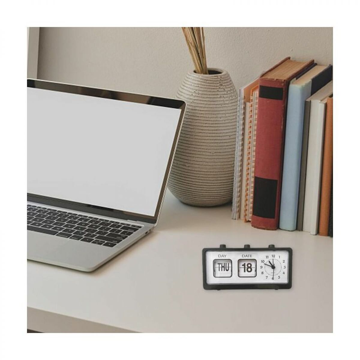 Ein Schreibtisch mit einem Laptop, einem Kalender-Uhr-Kombinationsgerät, einer dekorativen Vase mit Zweigen und mehreren aufrecht stehenden Büchern.