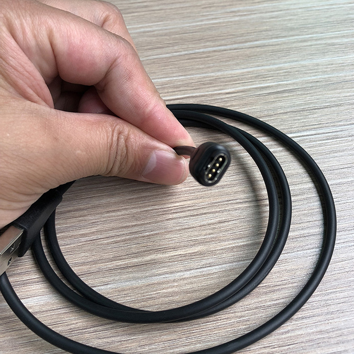Eine Hand hält einen schwarzen Ladekabel mit vier goldenen Pins vor einem Holzhintergrund.