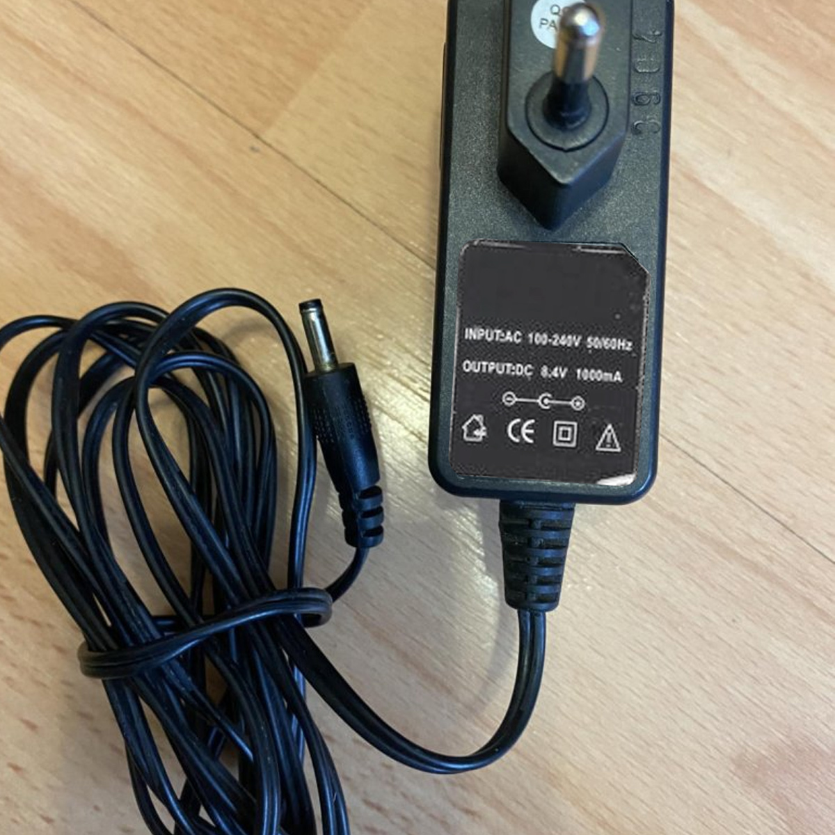 Ein schwarzes Netzteil mit einem EU-Stecker und einem langen, zusammengerollten Kabel liegt auf einem Holzfussboden.