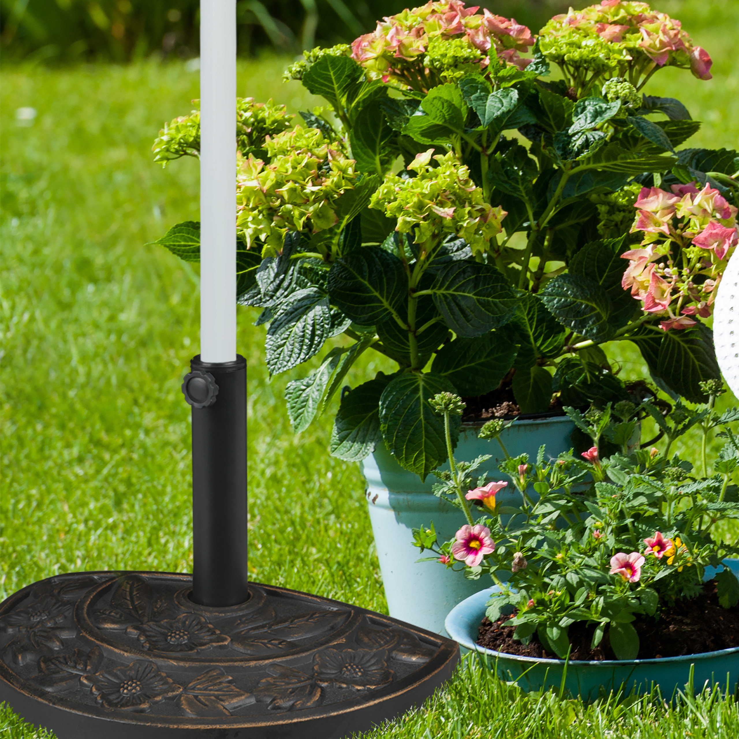 Ein dekorativer Sonnenschirmständer aus Gusseisen steht auf dem Rasen neben getopften Pflanzen und Blumen.