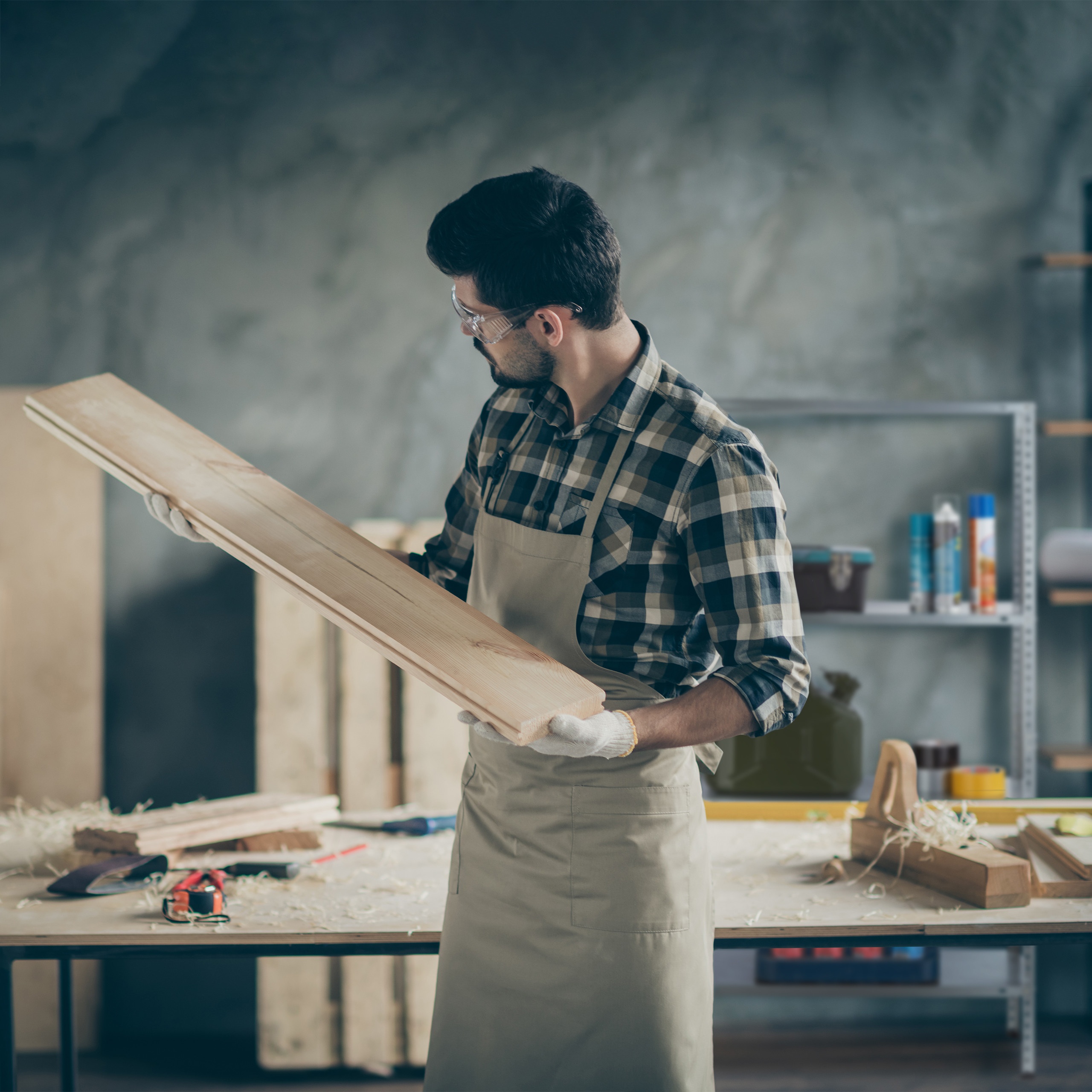 Ein Handwerker mit kariertem Hemd und Schürze inspiziert in einer Werkstatt ein langes Holzbrett.