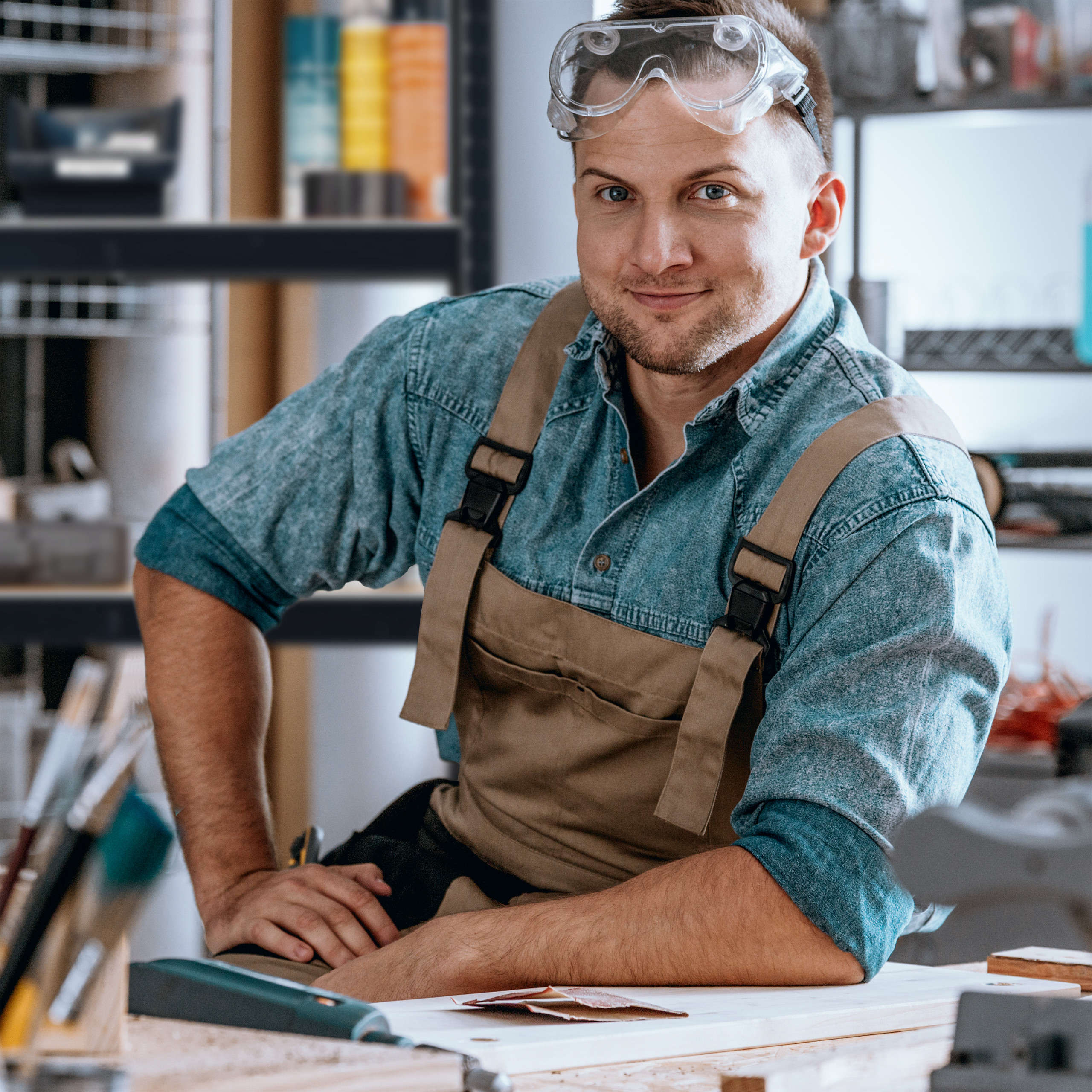 Ein lächelnder Handwerker in einer Werkstatt trägt eine Schutzbrille und eine braune Schürze.