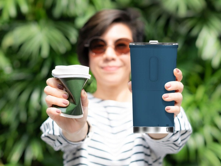 Eine Frau hält draußen einen zerknüllten Pappbecher in der einen und einen blauen Mehrwegbecher in der anderen Hand.