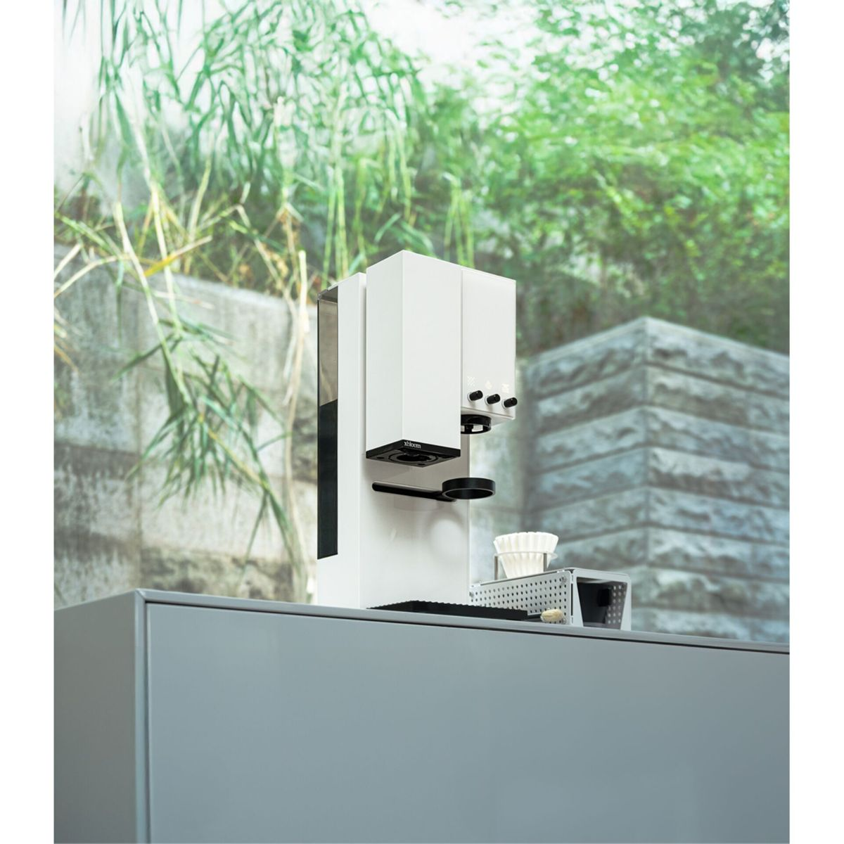 Eine weiße Kaffeemaschine steht auf einer Theke vor einer grünen Gartenwand.