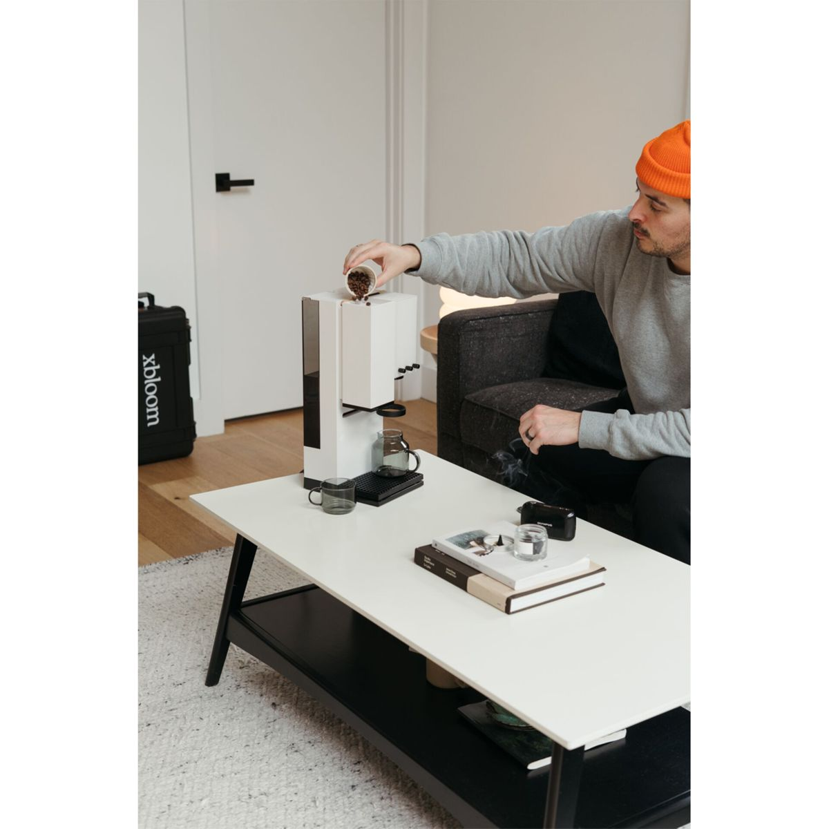 Ein Mann mit oranger Mütze gießt Kaffeebohnen in eine weiße Kaffeemaschine auf einem Couchtisch.