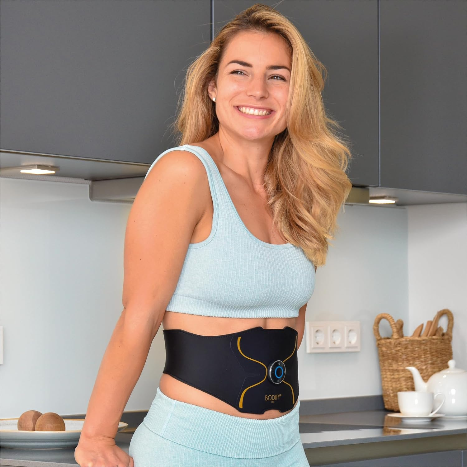 Eine lächelnde blonde Frau in Sportkleidung trägt einen schwarzen EMS-Bauchgurt in einer Küche.