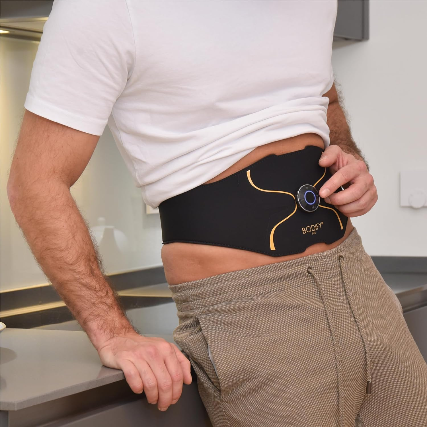 Ein Mann im weißen Shirt trägt einen schwarz-goldenen EMS-Trainingsgürtel am Bauch und drückt einen Knopf.