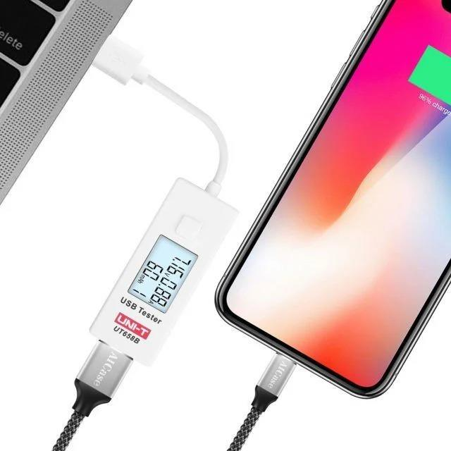 Bir dizüstü bilgisayar ile akıllı telefon arasında bağlı USB test cihazı, ekranda voltaj ve akım verilerini gösterir.