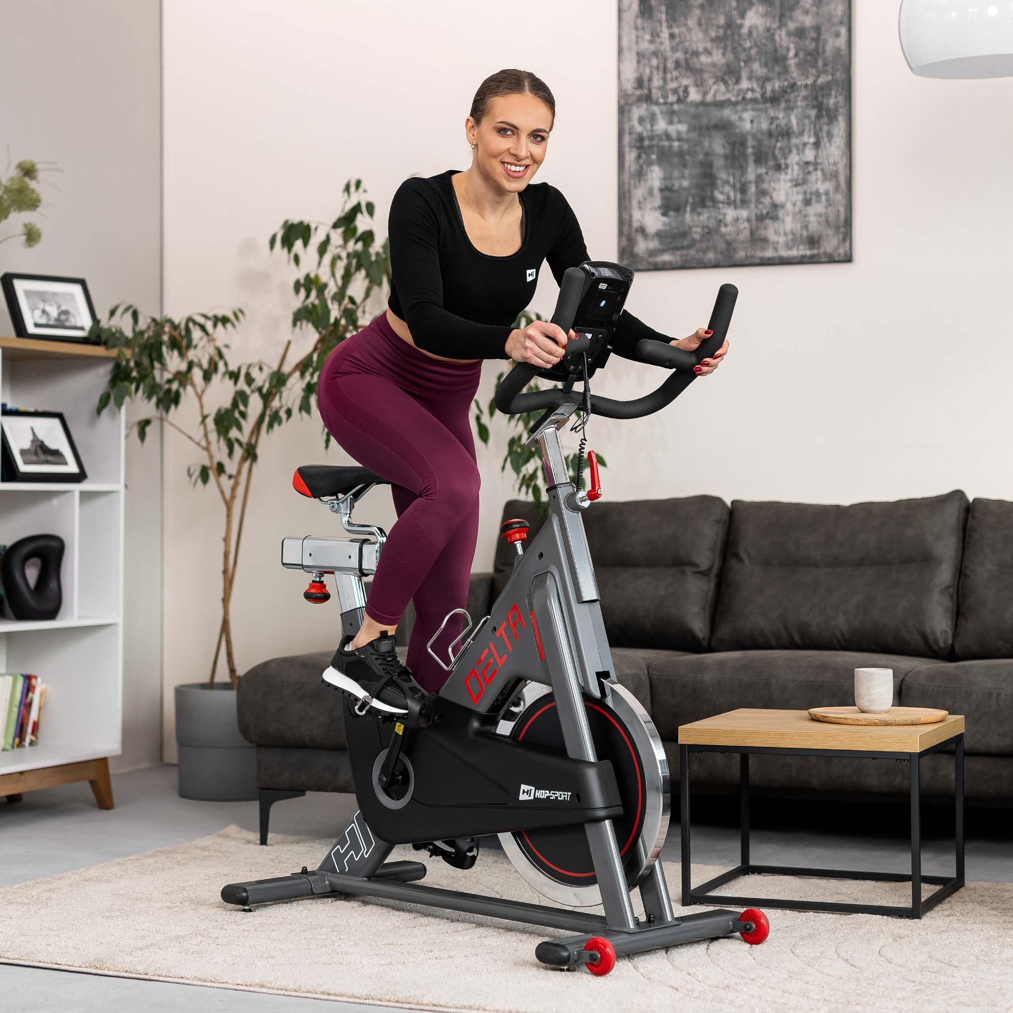 Eine lächelnde Frau in Sportkleidung trainiert auf einem grauen Heimtrainer in einem hellen Wohnzimmer.