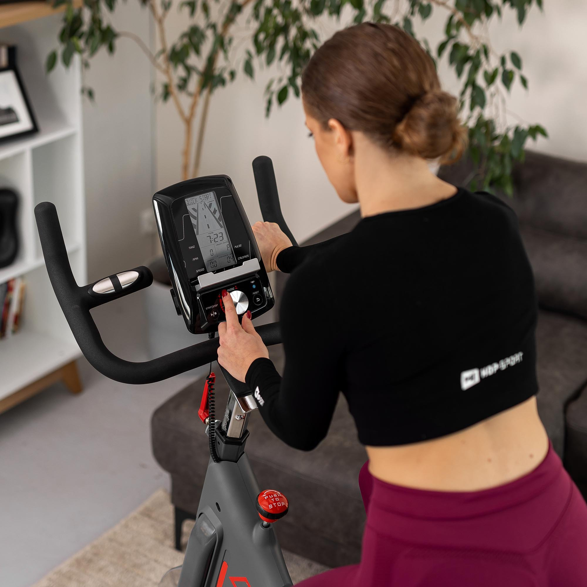 Eine Frau in schwarzem Oberteil und bordeauxroten Leggings trainiert drinnen auf einem Heimtrainer.