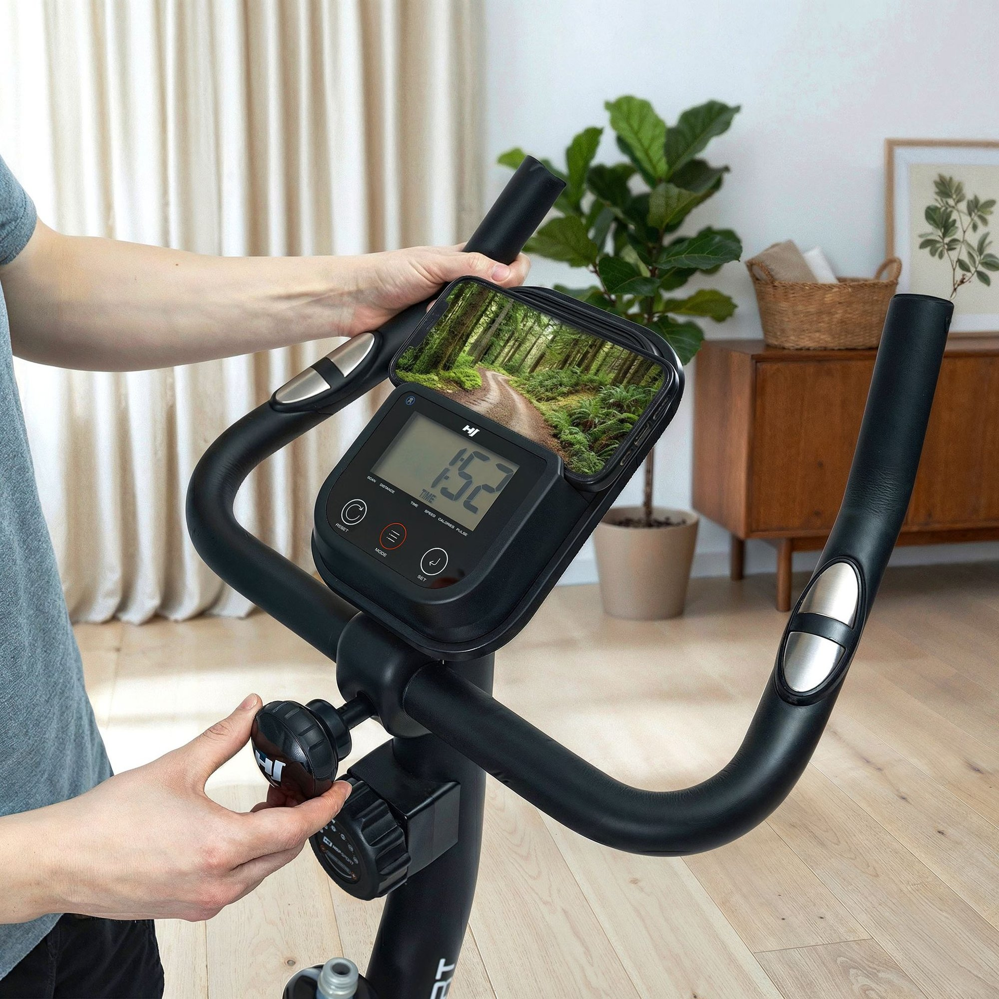 Person stellt ein schwarzes Heimfahrrad ein, mit Smartphone in Halterung und Digitaldisplay.