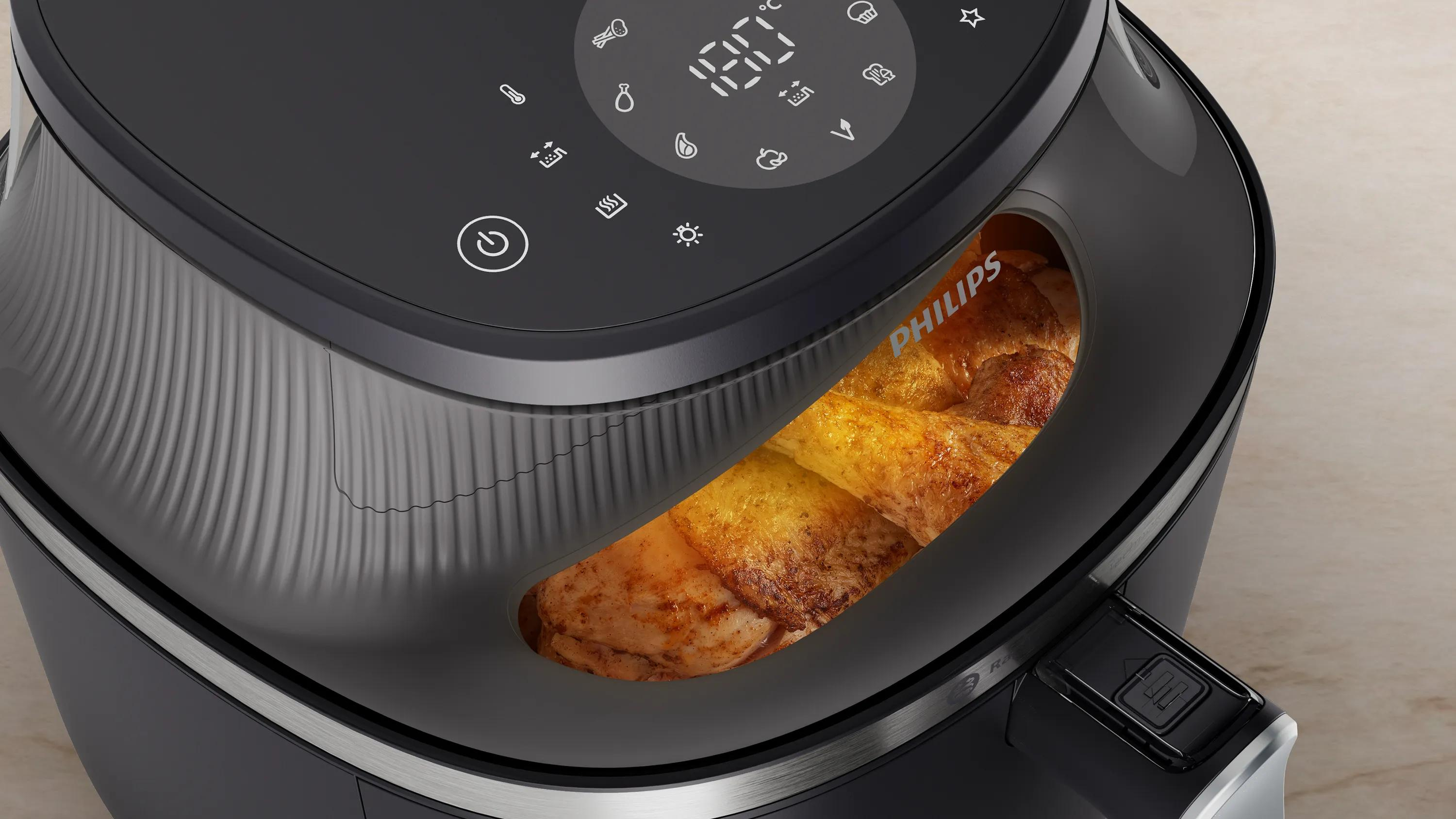 Una friggitrice ad aria Philips nera con display digitale mostra pezzi di pollo in cottura in un cestello illuminato.