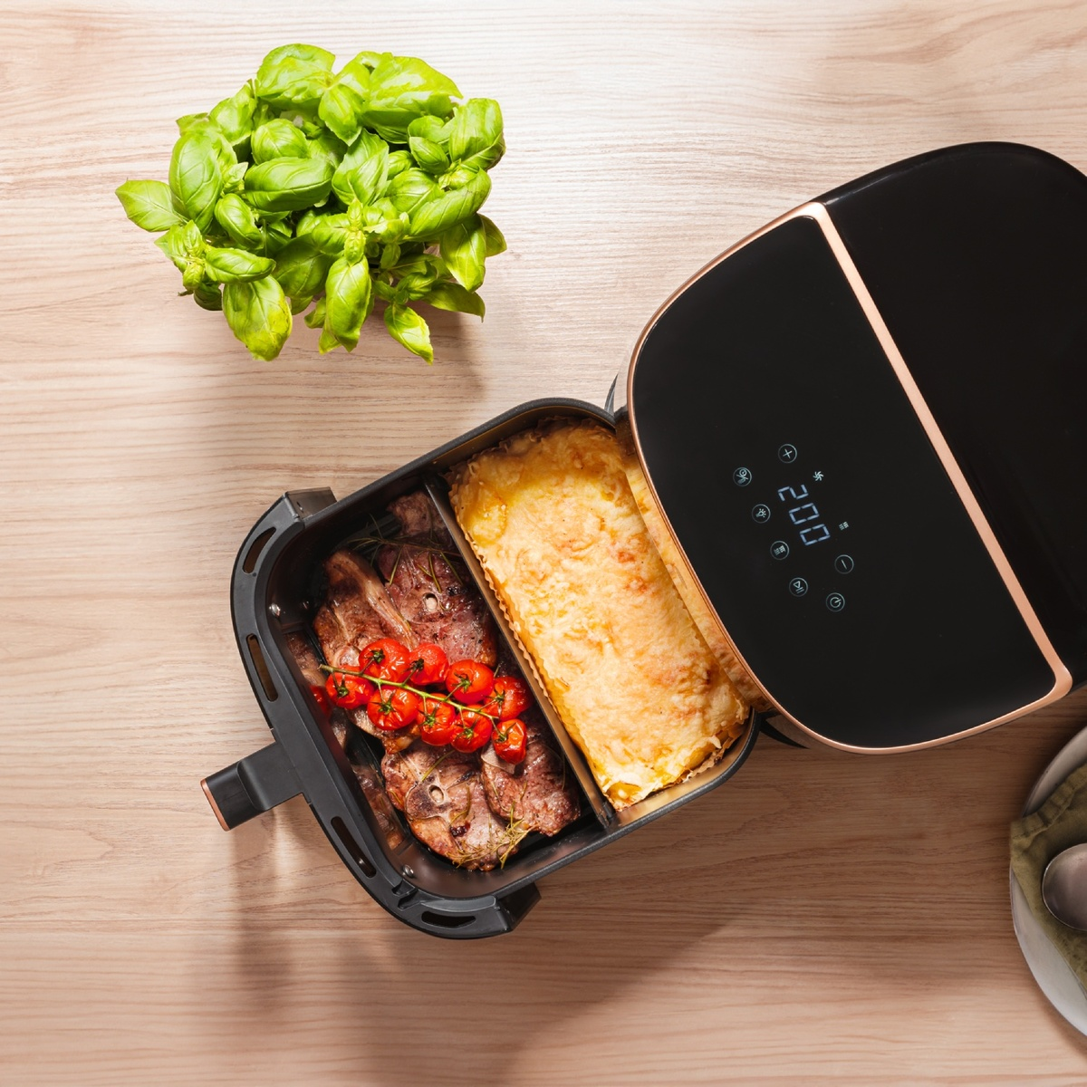 Eine offene schwarze Heißluftfritteuse mit Fleisch, Tomaten und Auflauf auf einem Holztisch, daneben frischer Basilikum.