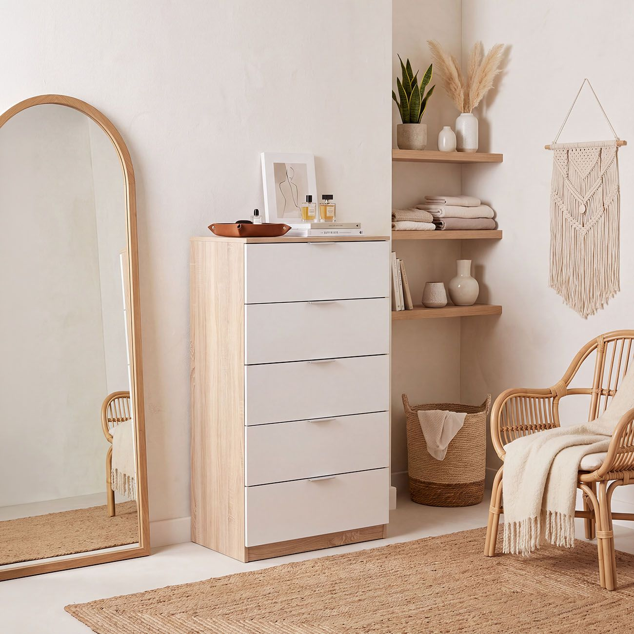 Una habitación con cómoda blanca, espejo de arco, silla de rattán, estantes con decoración y tapiz de macramé.