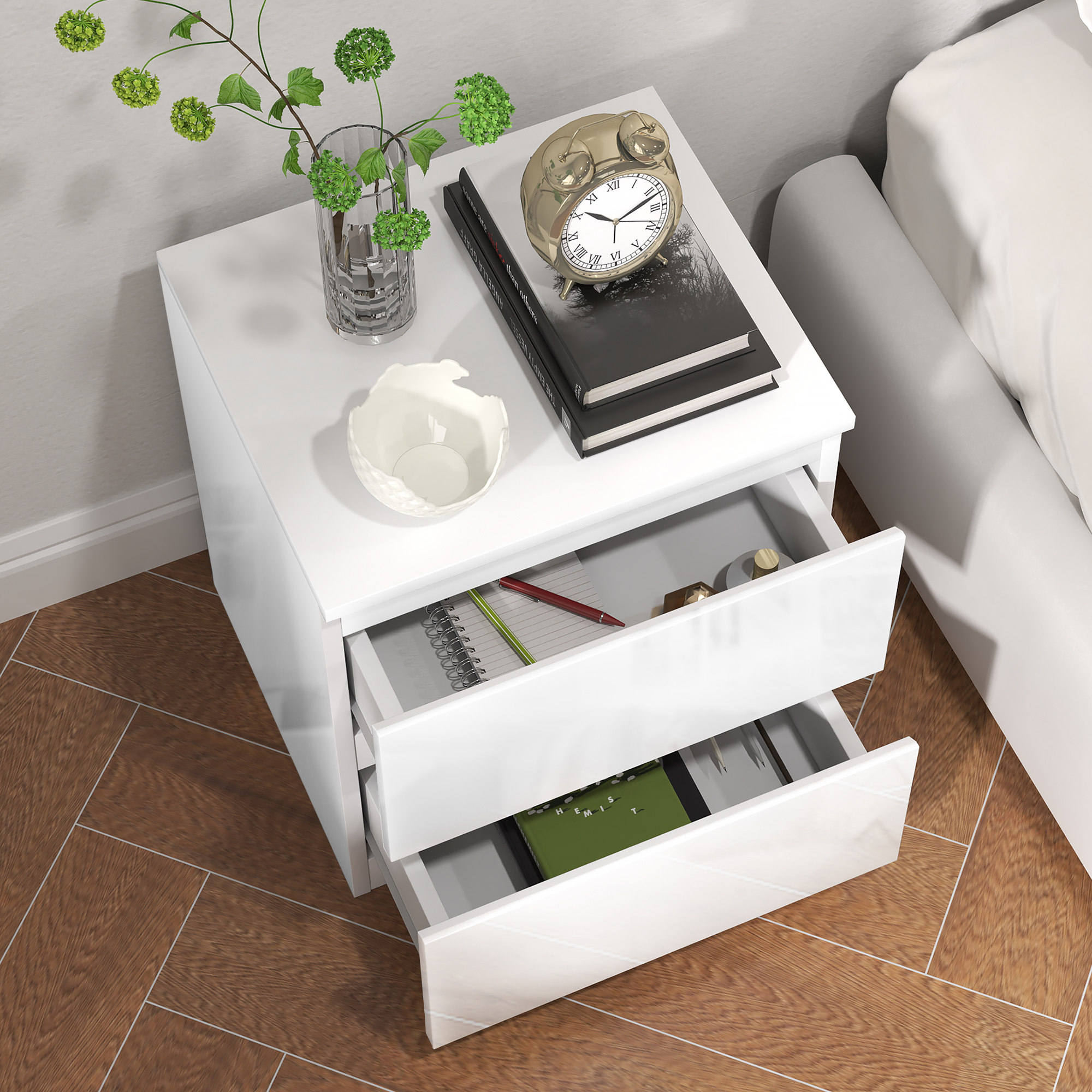 Mesita de noche blanca con cajones abiertos, jarrón, reloj y libros, al lado de una cama en suelo de madera.