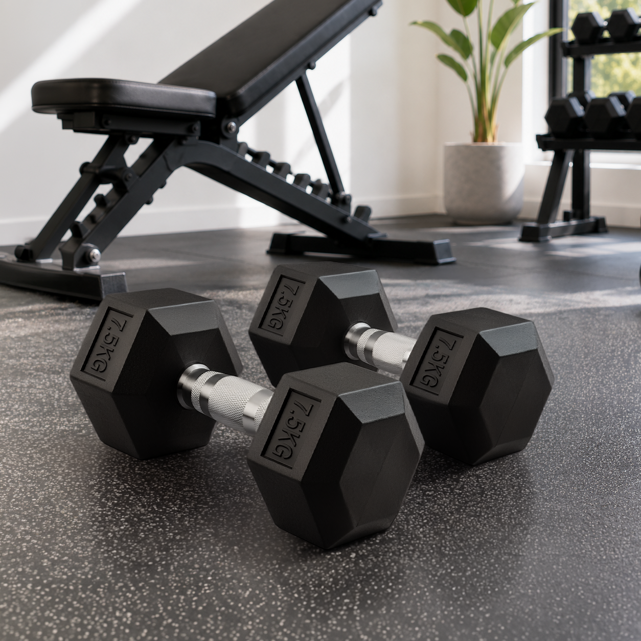 Dos mancuernas negras de 7,5 kg en el suelo del gimnasio, con un banco y un soporte al fondo.