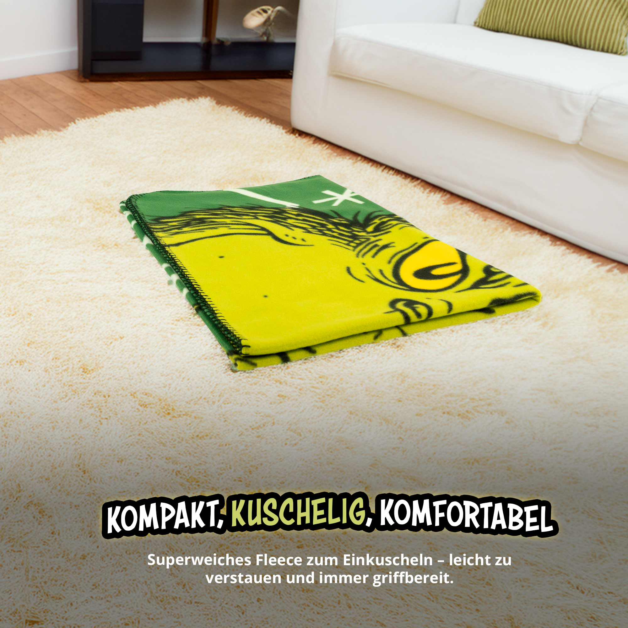 Eine gefaltete grün-gelbe Fleecedecke mit Grinch-Grafik liegt auf einem hellen Teppich vor einer weißen Couch.