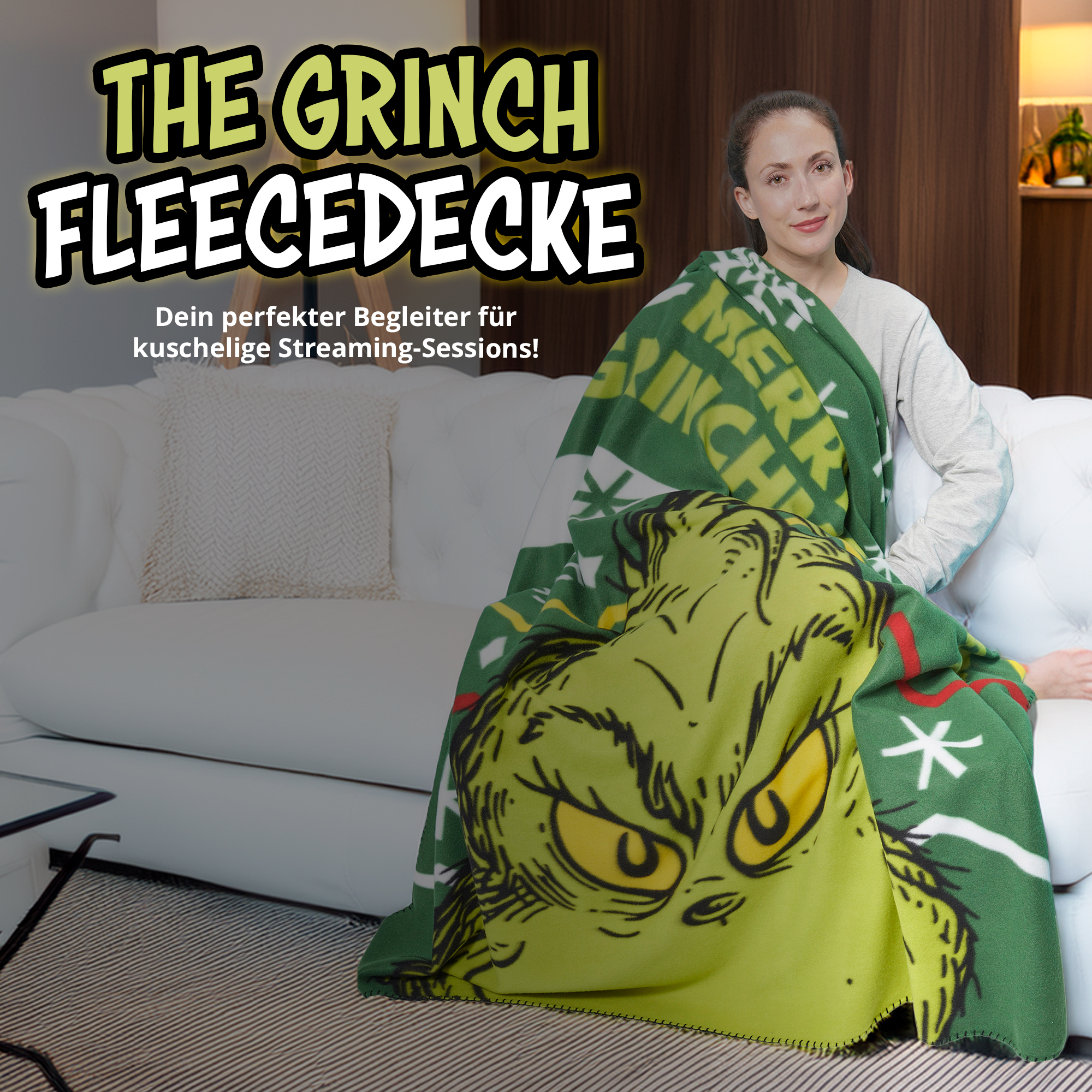 Eine Frau sitzt auf einer weißen Couch und ist in eine grüne Grinch-Fleececke gewickelt.