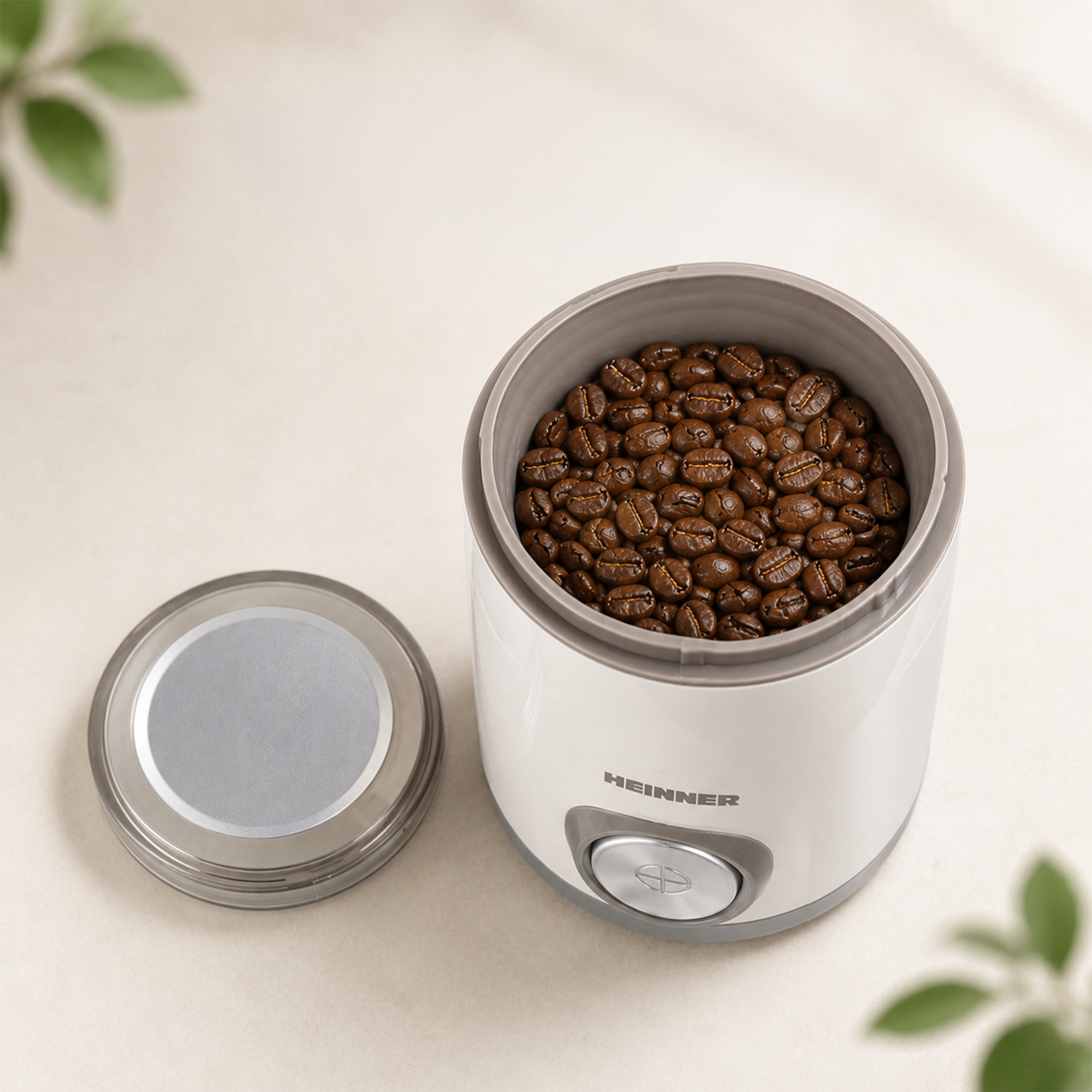 Eine offene weiße Kaffeemühle mit braunen Bohnen und abgenommenem Deckel steht auf einem hellen Tisch.