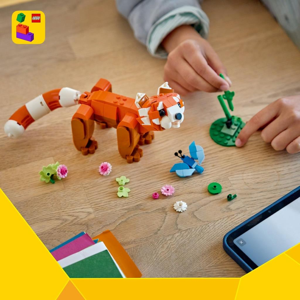 Bir çocuk, ahşap bir masa üzerinde küçük LEGO çiçekleri ve bir tabletle kırmızı bir panda LEGO modeli yapıyor.