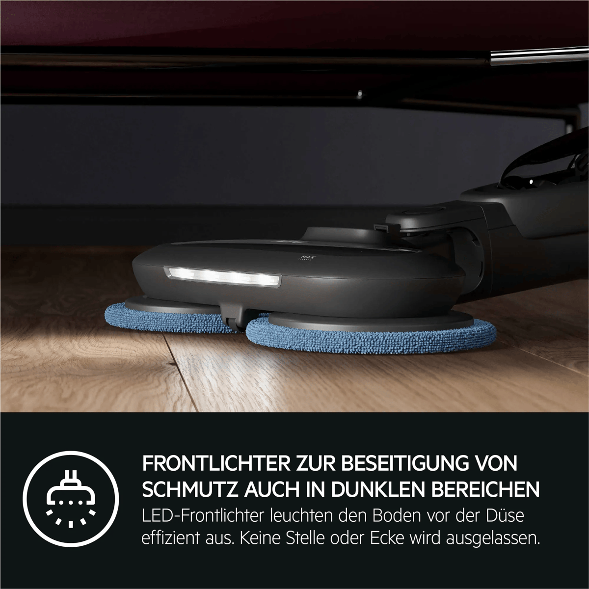 Una aspiradora mopa negra con luces LED frontales limpia el suelo de madera bajo un mueble.