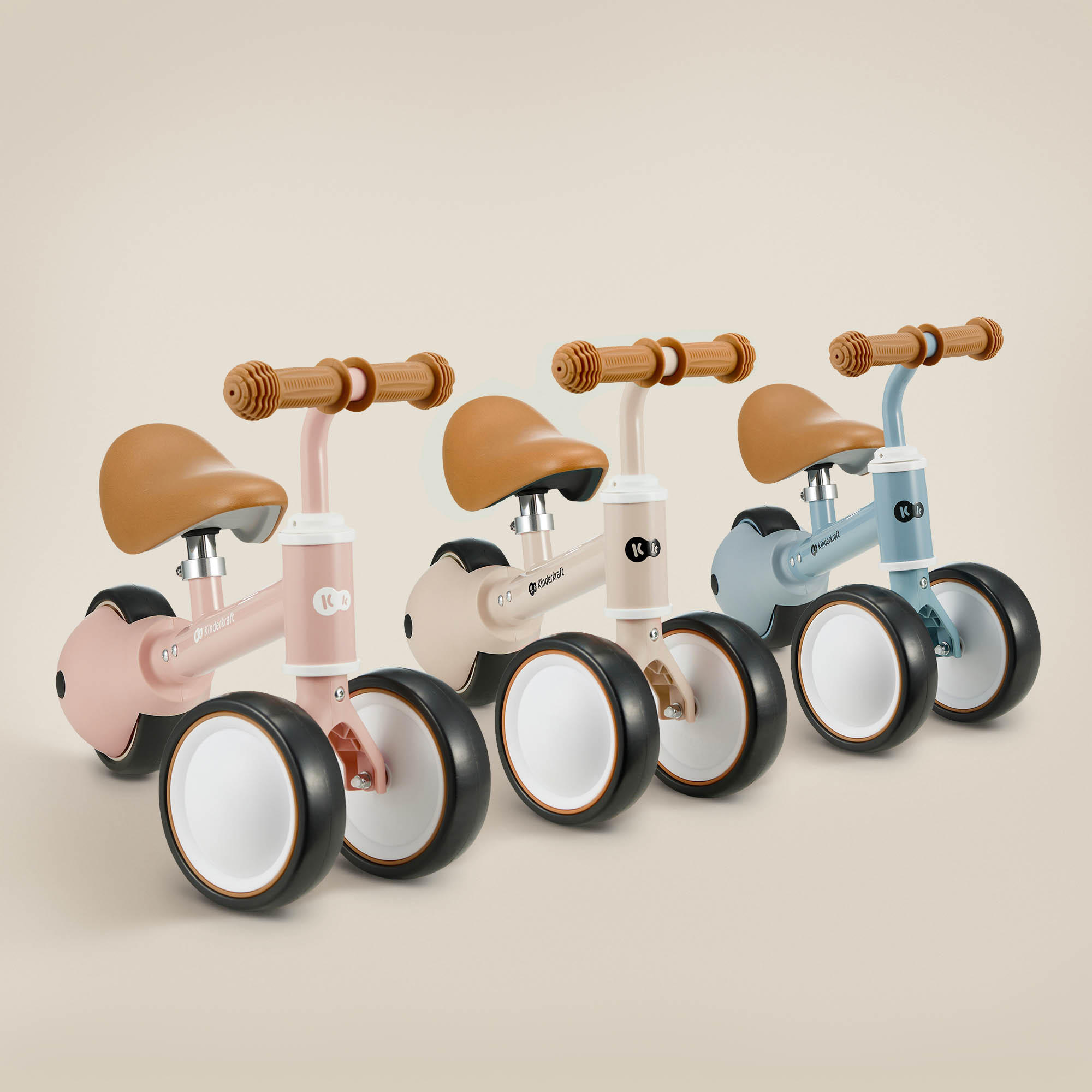 Drei kleine Laufräder in Rosa, Beige und Blau mit braunen Sätteln und Lenkern stehen vor neutralem Hintergrund.