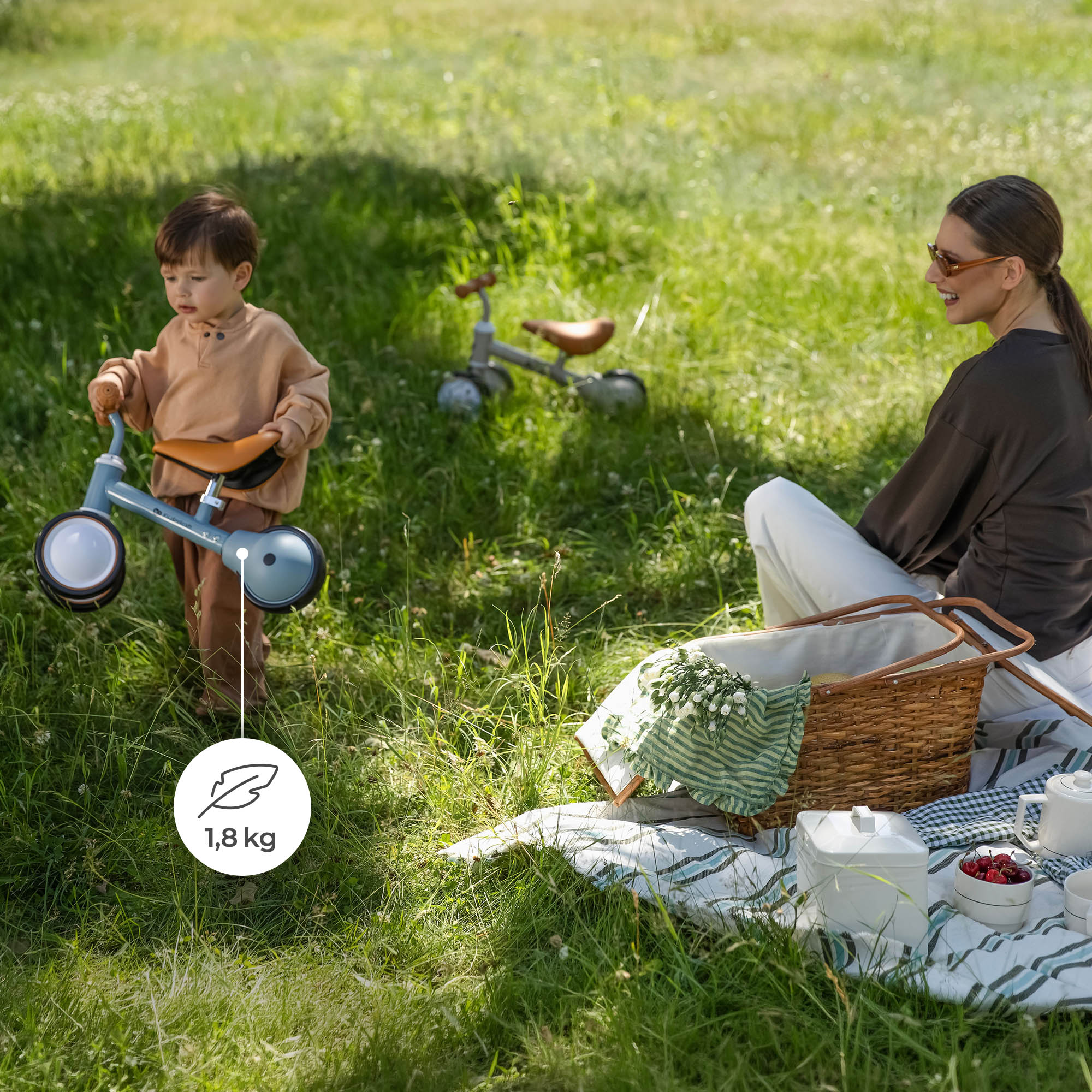 Kleinkind trägt ein leichtes blaues Laufrad auf einer Wiese, während die Mutter in der Nähe sitzt.