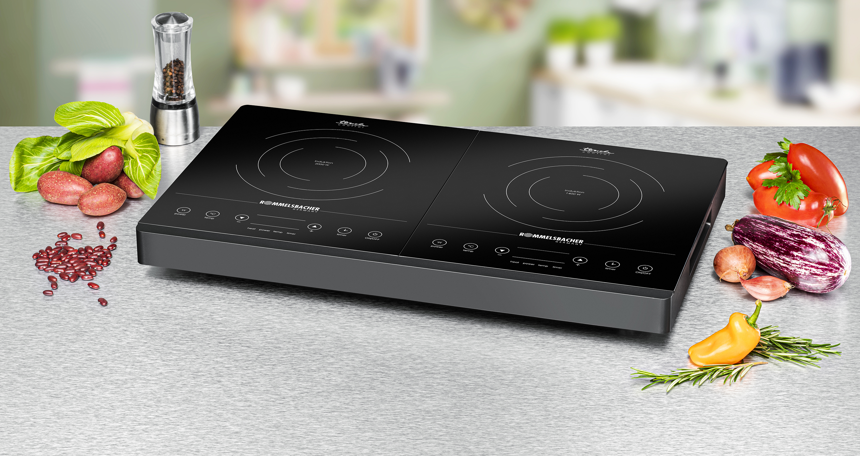 A black double induction cooktop on a stainless steel countertop, surrounded by fresh vegetables and spices.