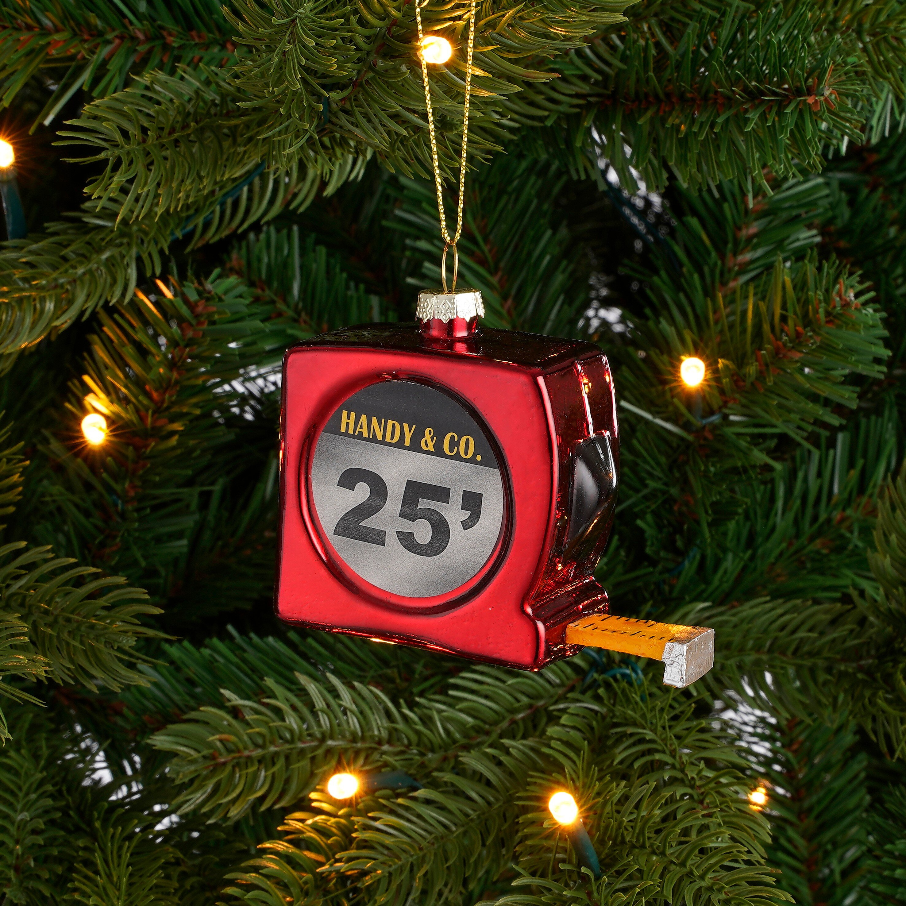 Een rood meetlint kerstboomornament hangt aan dennentakken met gloeiende lampjes.