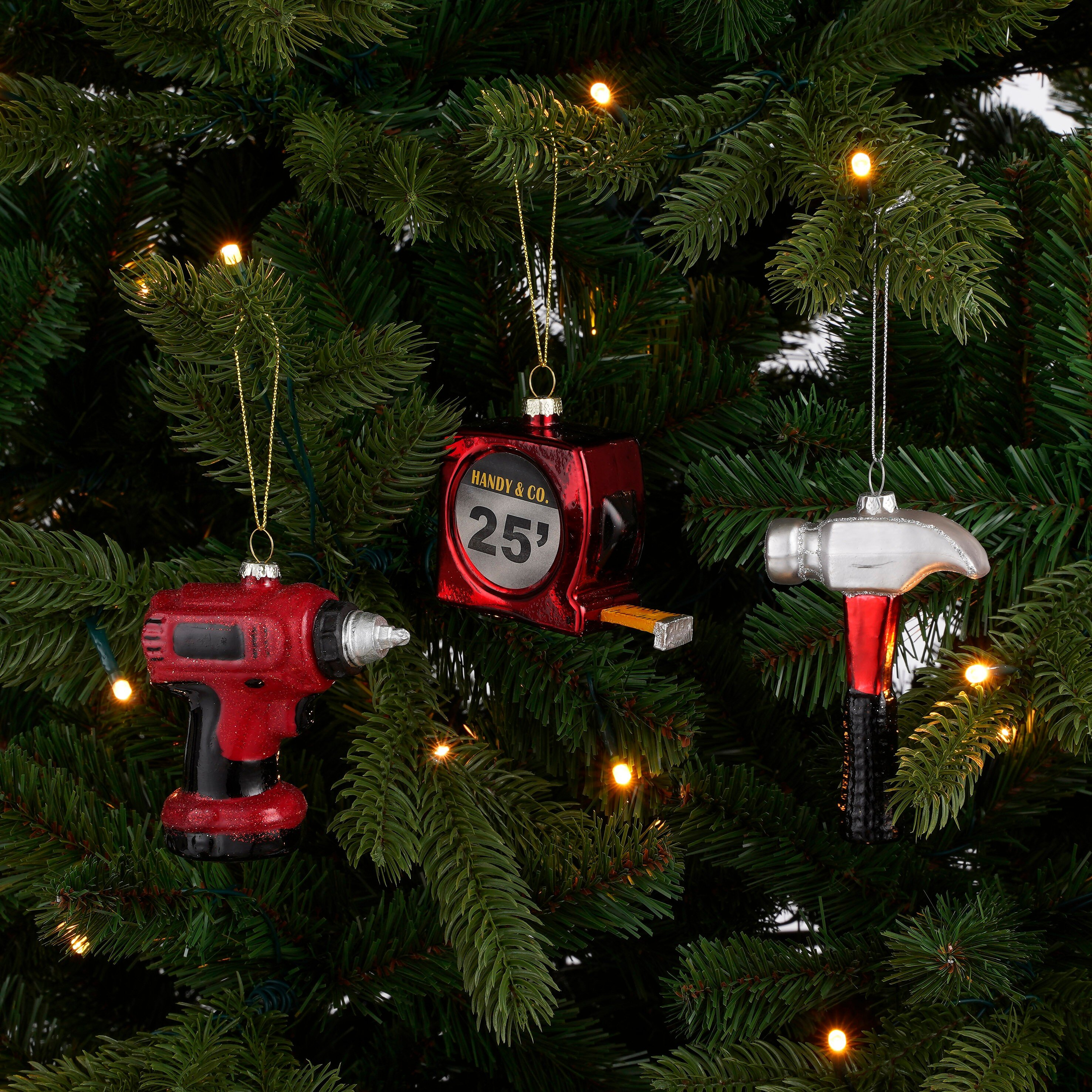 Drie gereedschapvormige kerstballen hangen in een groene kerstboom met lichtjes.