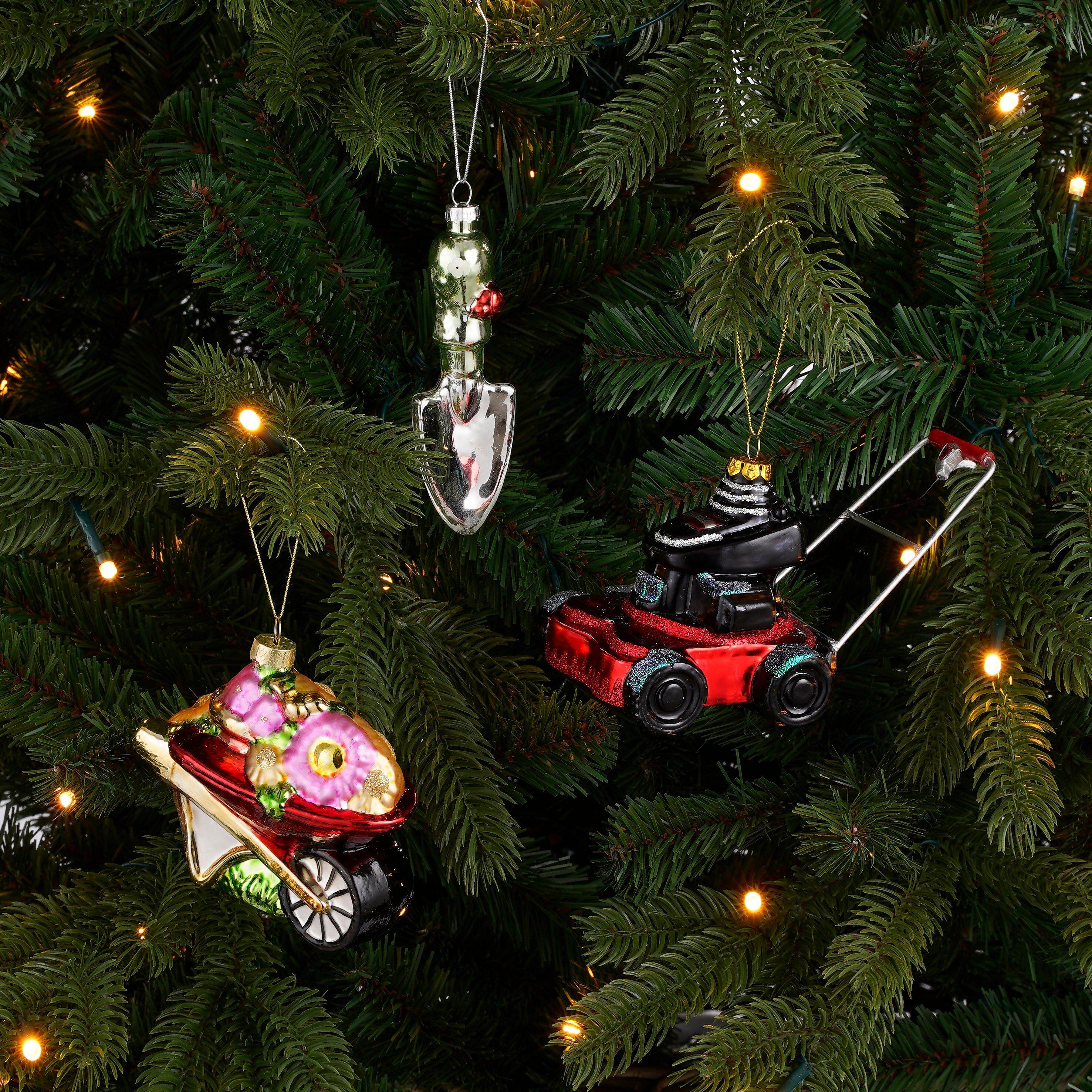Trois décorations de jardin sur un sapin illuminé : une pelle, une brouette fleurie et une tondeuse à gazon.