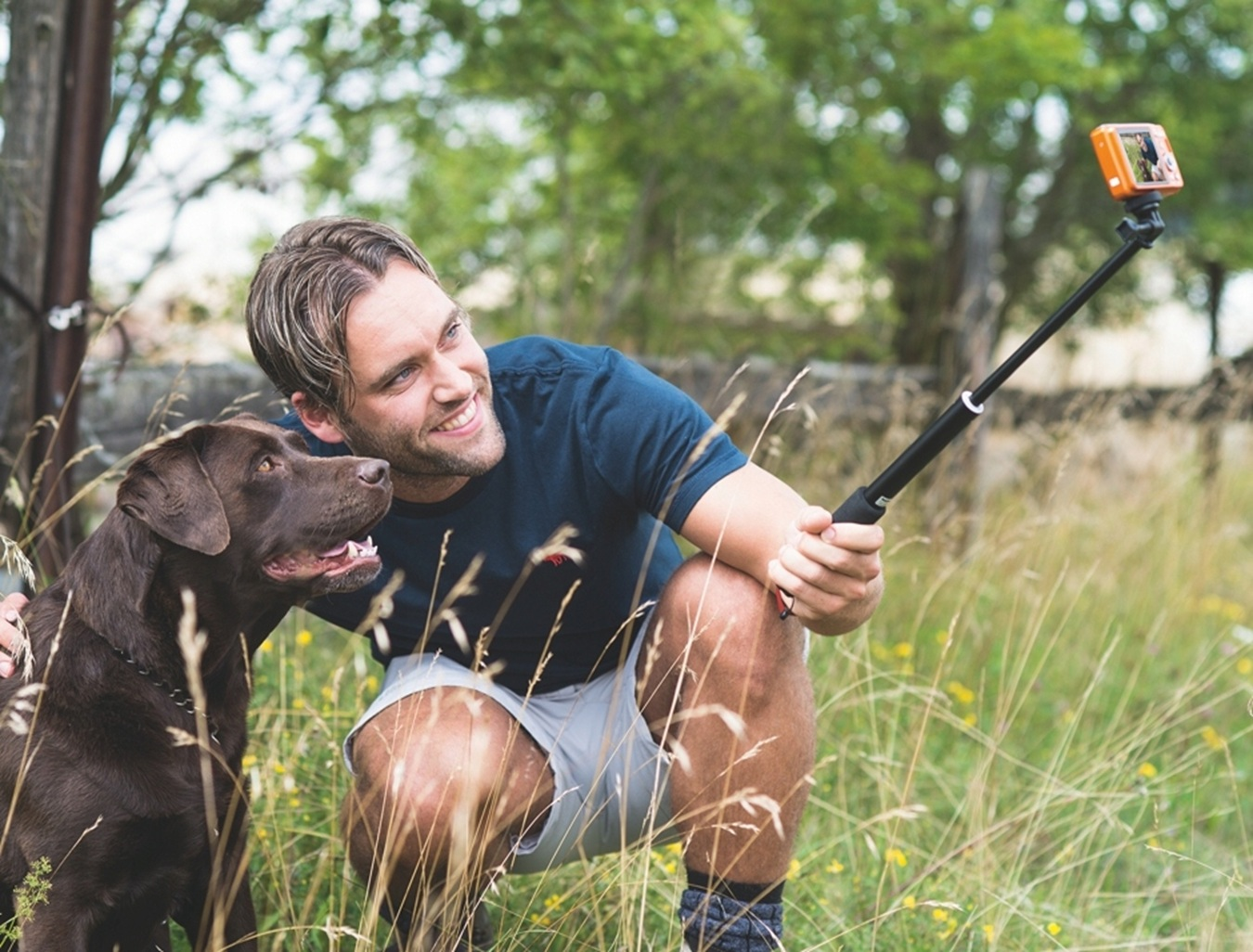 Un uomo sorridente e il suo labrador marrone accovacciati nell'erba alta fanno un selfie.