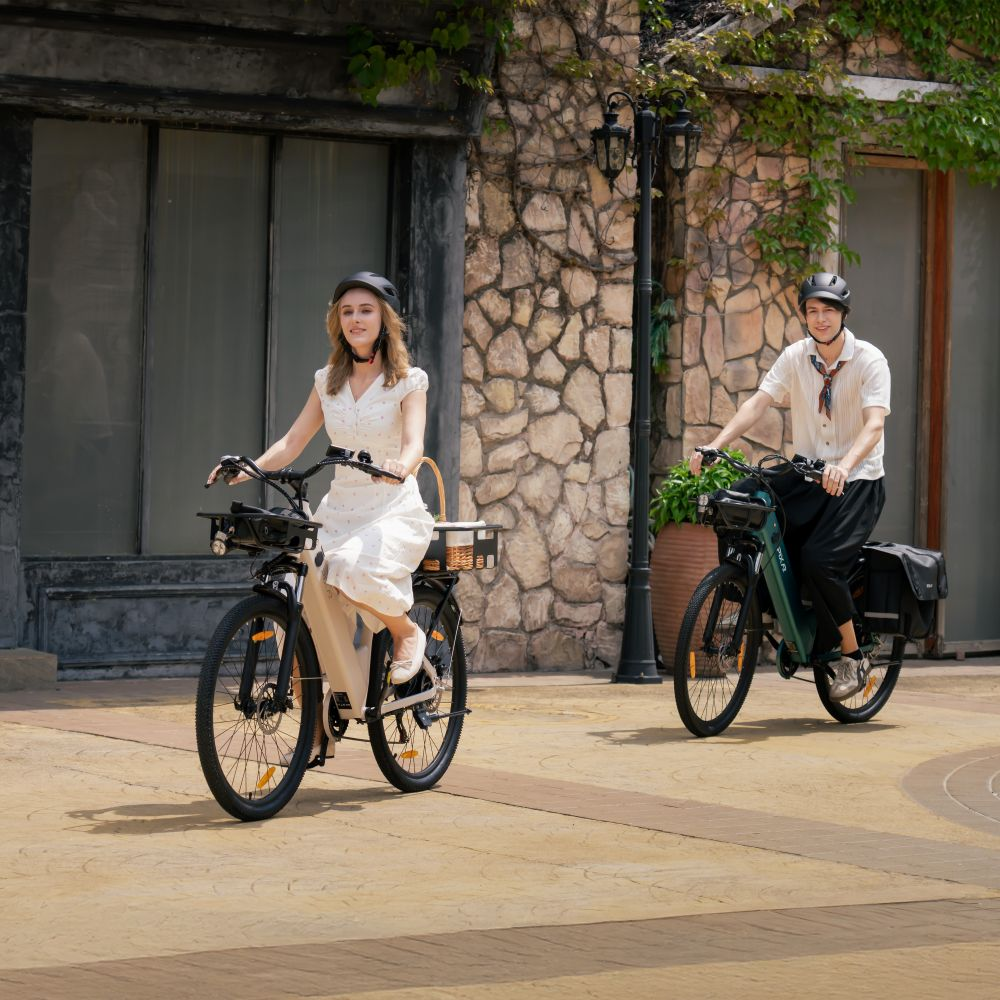 Una pareja monta en bicicleta eléctrica por una calle adoquinada frente a una casa de piedra con hiedra.