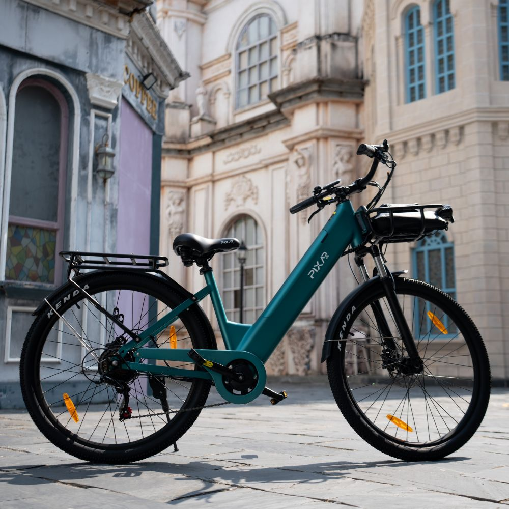 Una bicicleta eléctrica turquesa con cesta delantera y portaequipajes trasero aparcada sobre piedra frente a edificios.