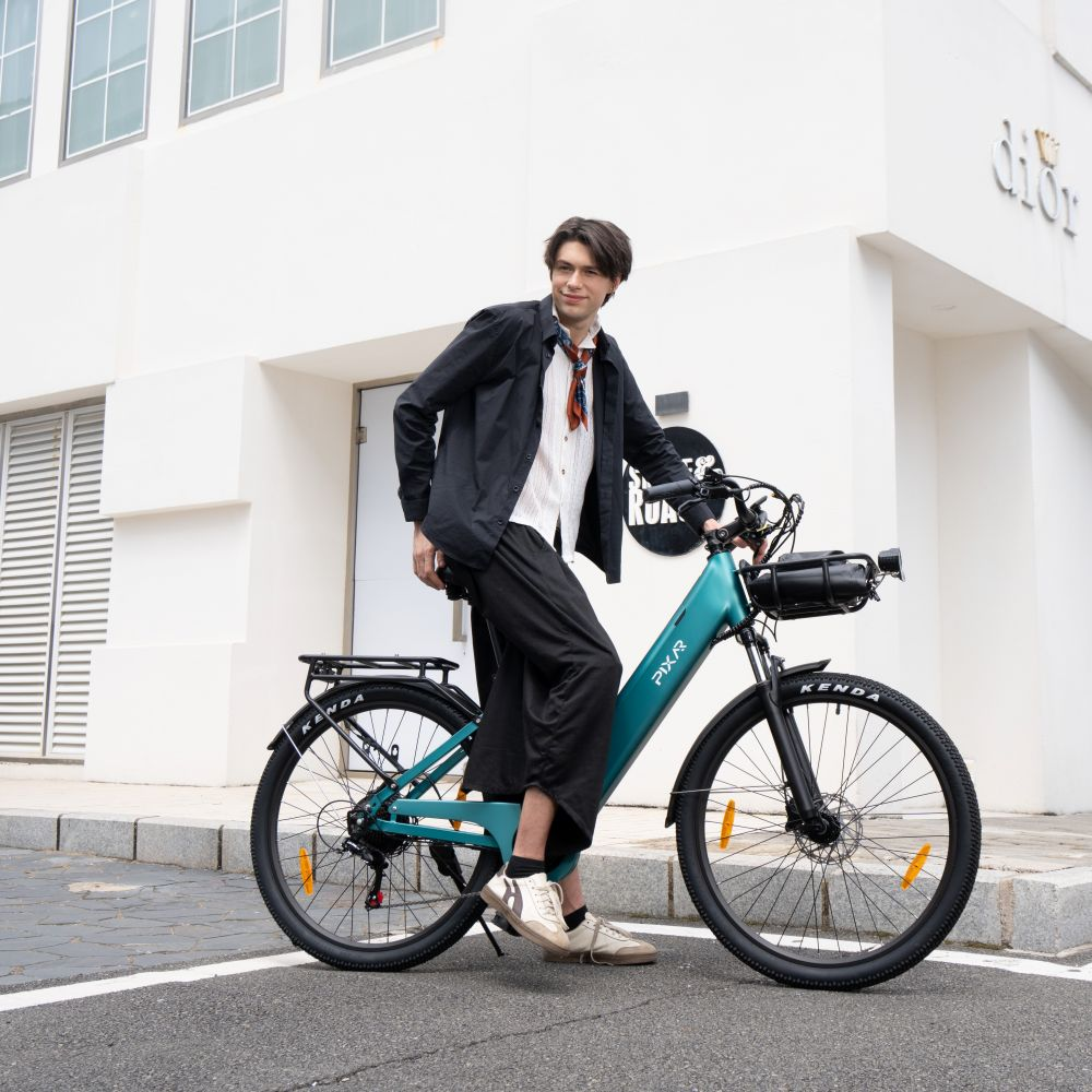 Un joven con camisa negra sonríe mientras posa en una bicicleta eléctrica verde frente a un edificio blanco.