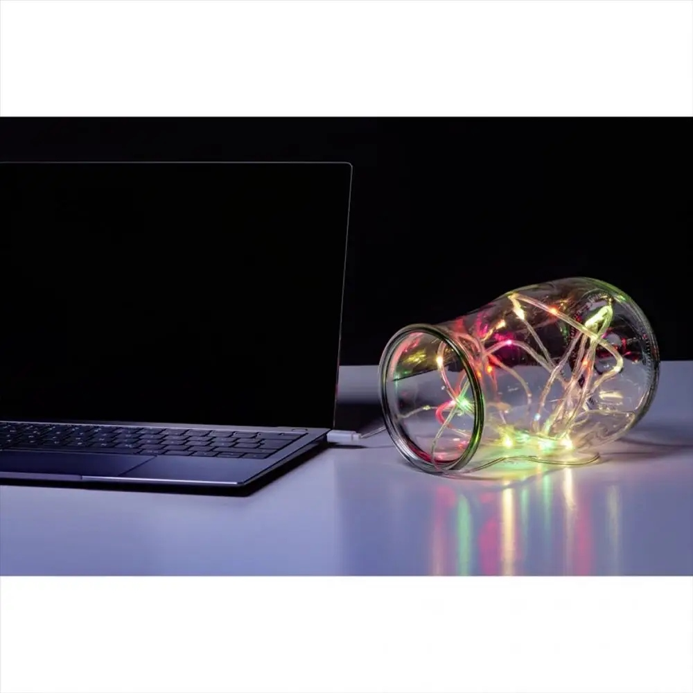 Laptop na ciemnej powierzchni obok szklanego słoika wypełnionego lampkami podłączonymi przez USB.