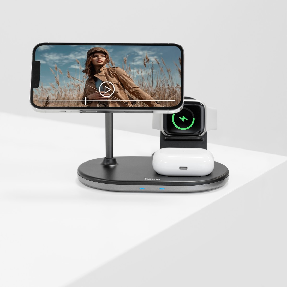 Czarna ładowarka bezprzewodowa trzyma smartfon, smartwatch i białe słuchawki na białym stole.