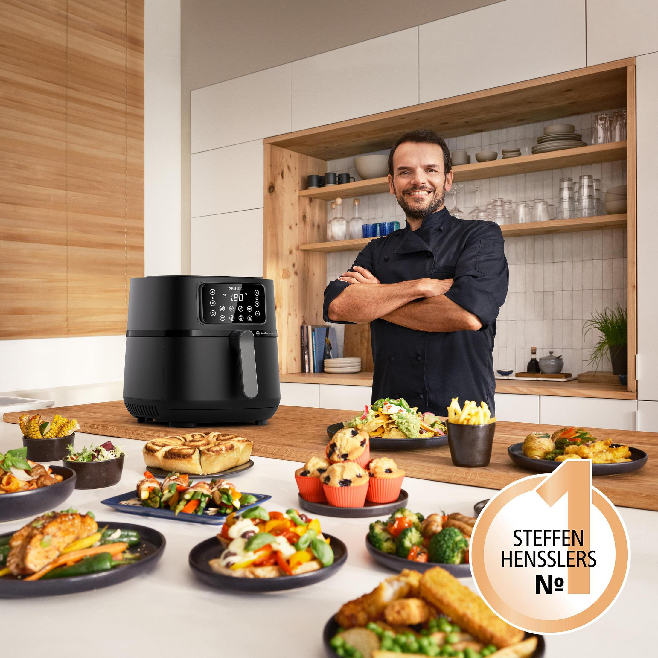 Uno chef si trova in una cucina con una friggitrice ad aria nera e vari piatti sui ripiani di legno.