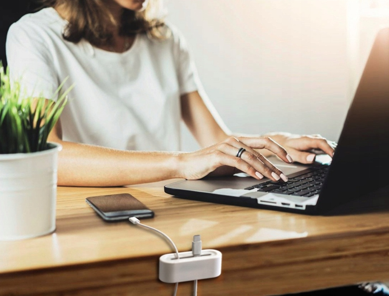 Kobieta pisze na laptopie przy drewnianym biurku, na pierwszym planie ładowany smartfon i uchwyt na kabel.