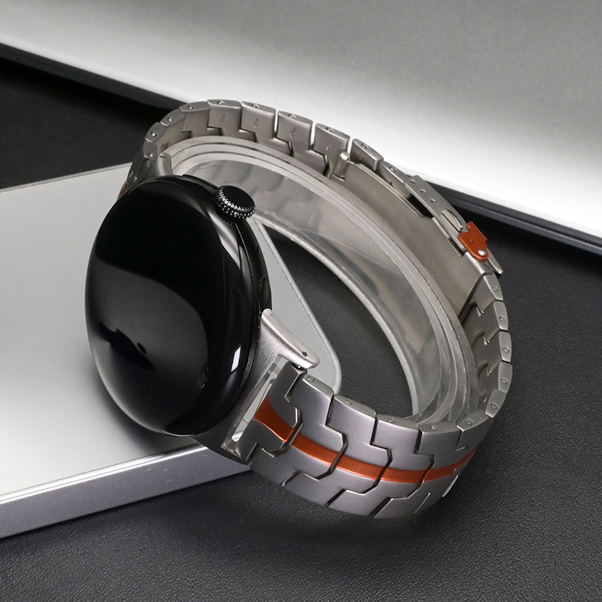 Eine schwarze Smartwatch mit silber-orangefarbenem Gliederarmband liegt auf einer Metalloberfläche.