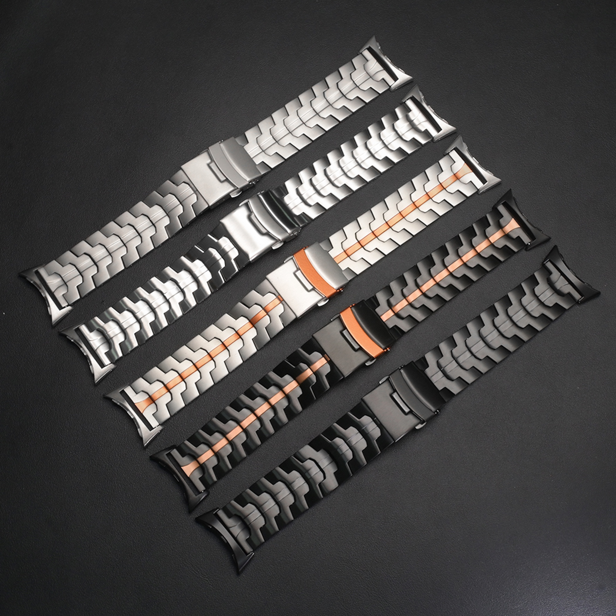 Fünf verschiedene Metall-Uhrenarmbänder in Silber, Schwarz und mit roségoldenen Akzenten auf dunklem Hintergrund.