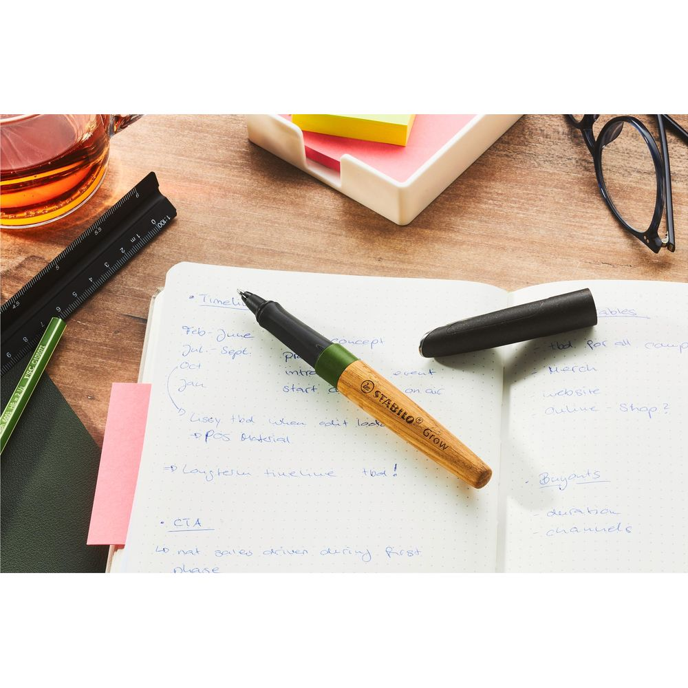 Ein Stabilo-Stift mit Holzschaft auf einem Notizbuch mit handgeschriebenen Notizen und Büroartikeln auf Holztisch.